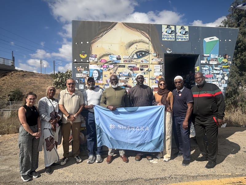 Sharaka delegation holding the Sharaka banner in front of a memorial mural