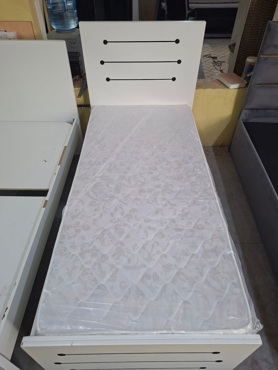 Top-down view of used white single bed frame with floral-pattern mattress still in plastic wrap