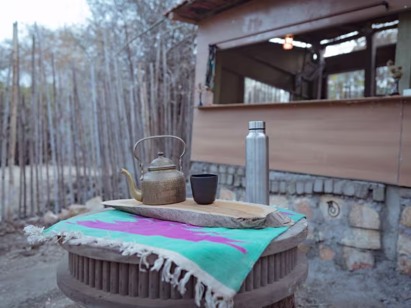 A morning chai setup outside The Pods at Limban — a brass teapot, steel flask and small cup arranged on a traditional round stone table, with a folded textile and bamboo grove in the background.