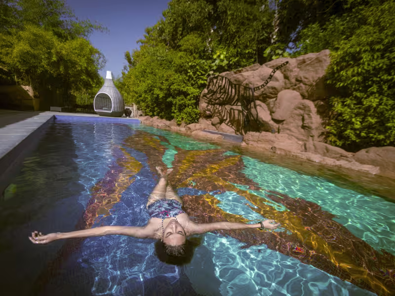 A female guest relaxing in Limban's 25-metre outdoor swimming pool, with natural stone surrounds, an adjoining kids' pool and dense greenery in the background.