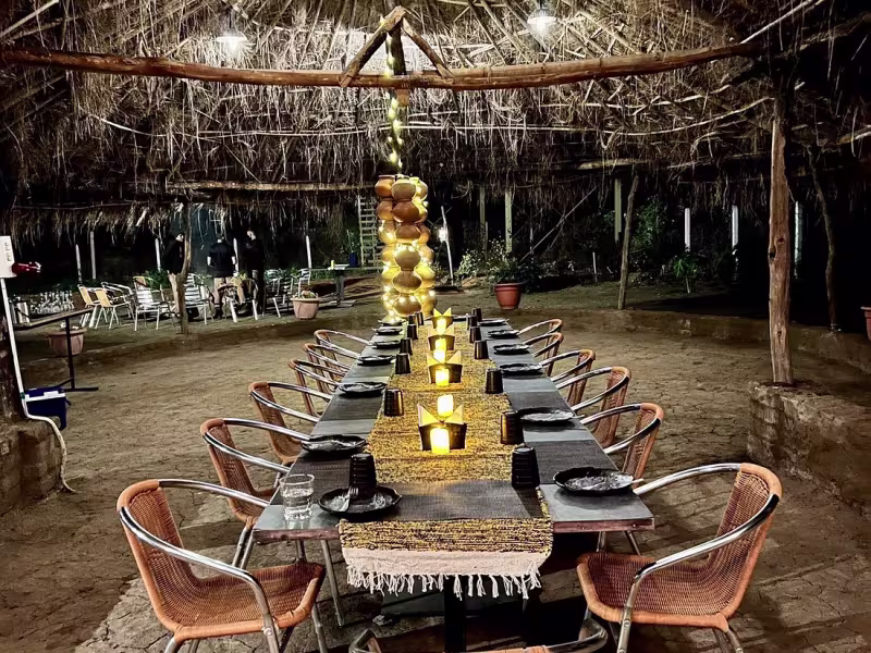 Special occasion long-table dinner setup under a bamboo-and-thatch canopy at Limkheda, Limban's open-air event ground, with candles, woven chairs and warm golden lighting at night.