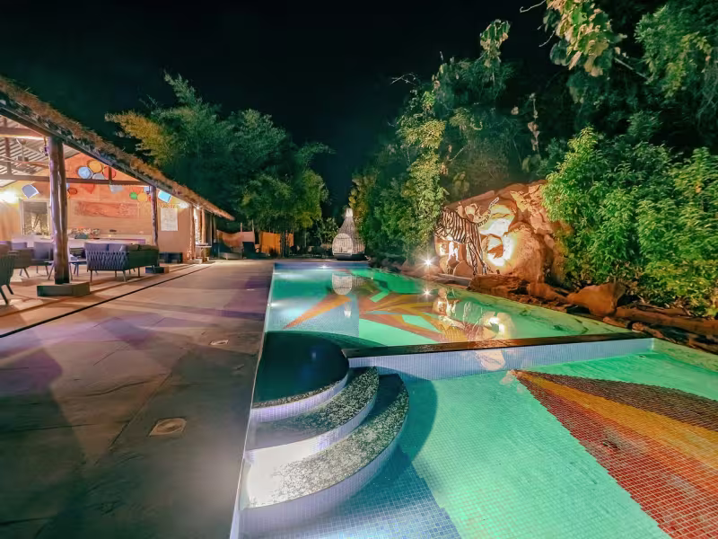 Wide-angle night view of Limban's 25-metre outdoor swimming pool illuminated in blue-green, with an adjoining kids' pool, sculpted rocks and tree cover around the perimeter.