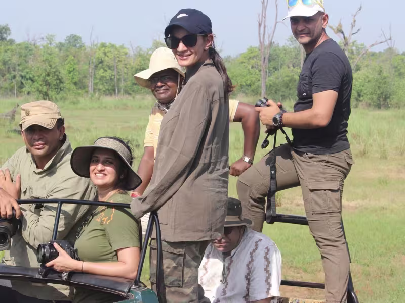 Six guests seated and standing in an open Limban safari jeep in a grassland clearing, equipped with cameras, caps and binoculars, heading out for an afternoon tiger safari in Tadoba.