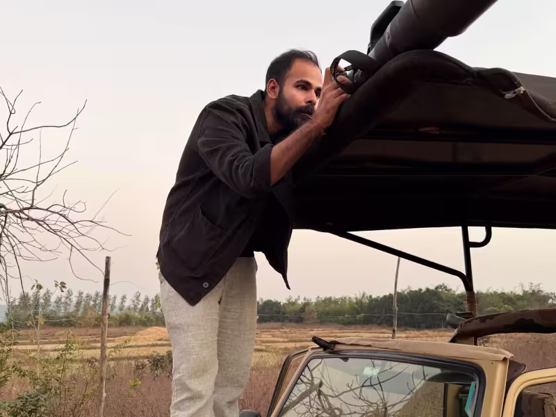 A male guest standing on the bonnet of a Limban safari jeep, peering through a large telephoto lens to photograph birds in Tadoba's open grassland at dawn.