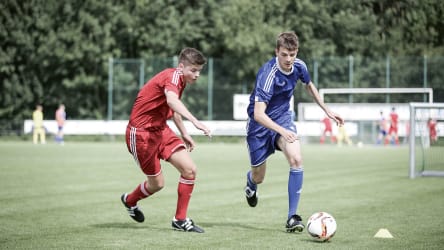 Zwei jugendliche Fußballer im Zweikampf um den Ball © 2019 Philippka-Sportverlag Zwei jugendliche Fußballer im Zweikampf um den Ball. Gespielt wird auf Kunstrasen und während der Junge im blauen Trikot versucht, am Gegenspieler in Rot vorbei zu laufen, attackiert dieser den ballführenden Spieler © 2019 Philippka-Sportverlag