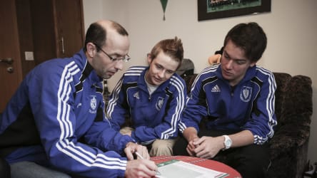 Die Spieler und der Trainer erstellen einen Wochenplan © 2019 Philippka-Sportverlag Der Trainer sitzt mit seinen Spielern an einem Couchtisch und beredet den Wochenplan. Alle gucen auf das Blattpapiert und der Trainer schreibt mit einem Stift darauf. © 2019 Philippka-Sportverlag
