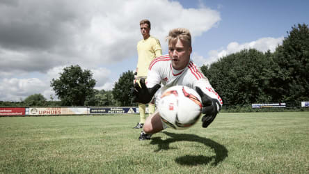 Der Torwart hat den Ball fest im Blick © 2019 Philippka-Sportverlag Ein Ball ist im Zentrum des Bildes und ist kruz davor von einem Torwart gefangen zu werden. Sein Trainer steht daneben und bep´obachtet die Ausfürhung der Übung. © 2019 Philippka-Sportverlag