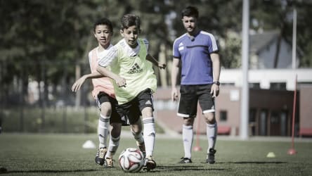 Spielerisch wird ein technisch-tatktisches Fundament gelegt © 2019 Philippka-Sportverlag Ein junger Spieler dribbelt mit dem Ball seinem Traingspartner davon, während der Trainer von hinten alles beobachtet. Im Hintergrund ist ein kleiner Dribbel-Slalom aus Stangen zu sehen.© 2019 Philippka-Sportverlag