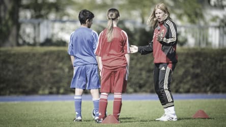 Die Trainerin gibt den Spieler direkt nach der Übung Tips © 2019 Philippka-Sportverlag Ein Junge und ein Mädchen stehen auf dem Platz vor ihrer Traininerin und hören ihr zu, was sie bei der Übung noch verbessern können. Auf dem Feld sind rote Hütchen zu sehen. © 2019 Philippka-Sportverlag