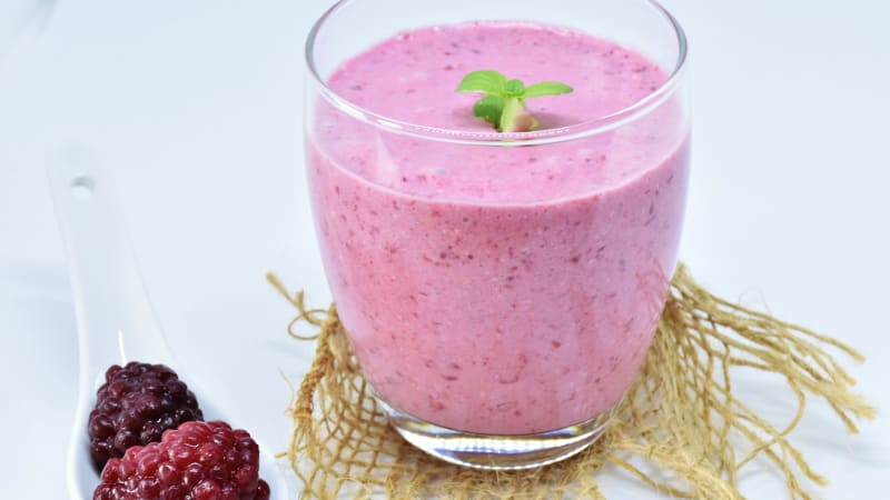 Das Bild zeigt ein Glas mit einem Rosafarbenen Kokusshake mit Beeren. Neben dem Glas ist ein weißer Keramik Löffel auf welchem Himbeeren liegen.