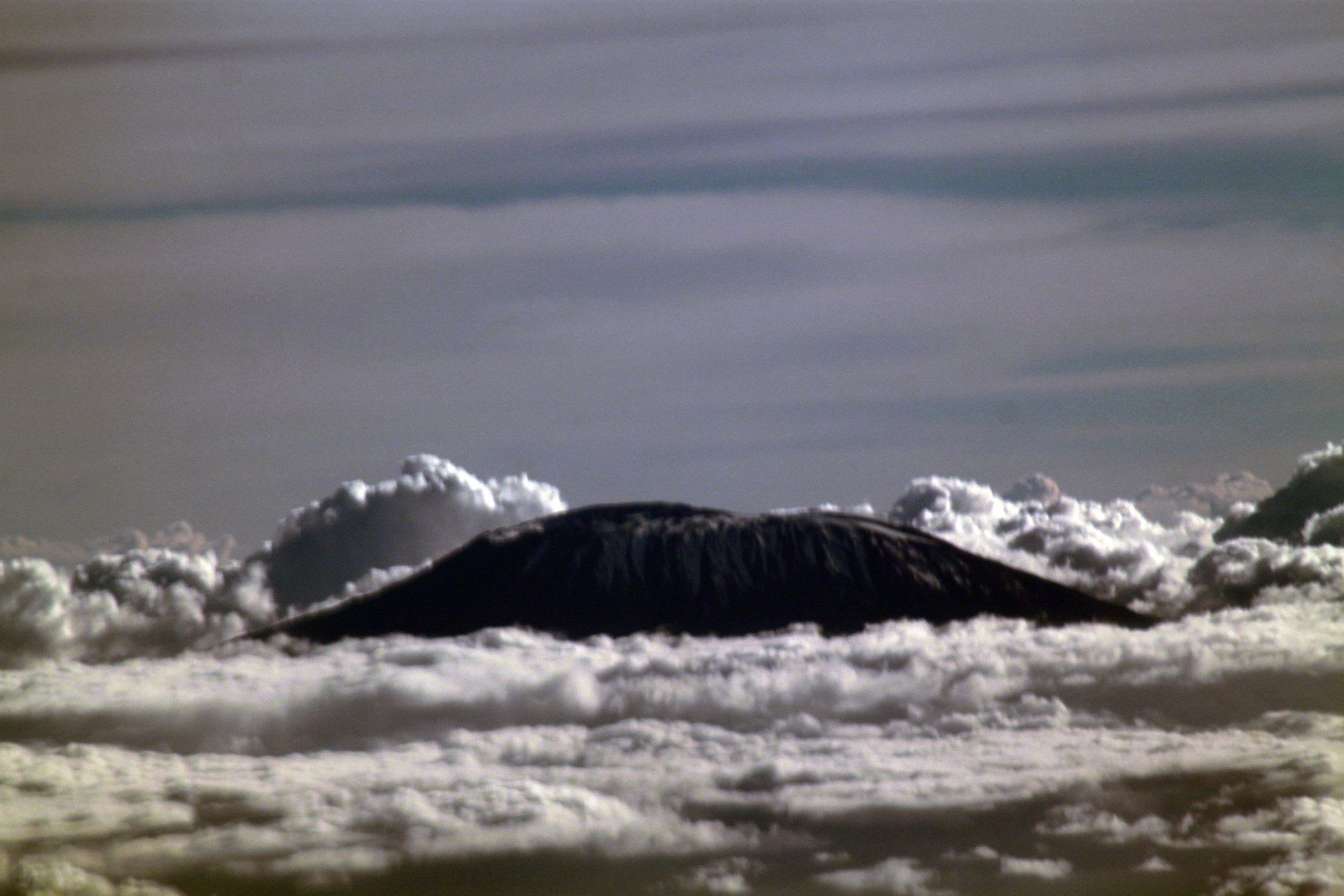 Dark, rounded mountain summit rising above a sea of white cloud under a streaked grey sky