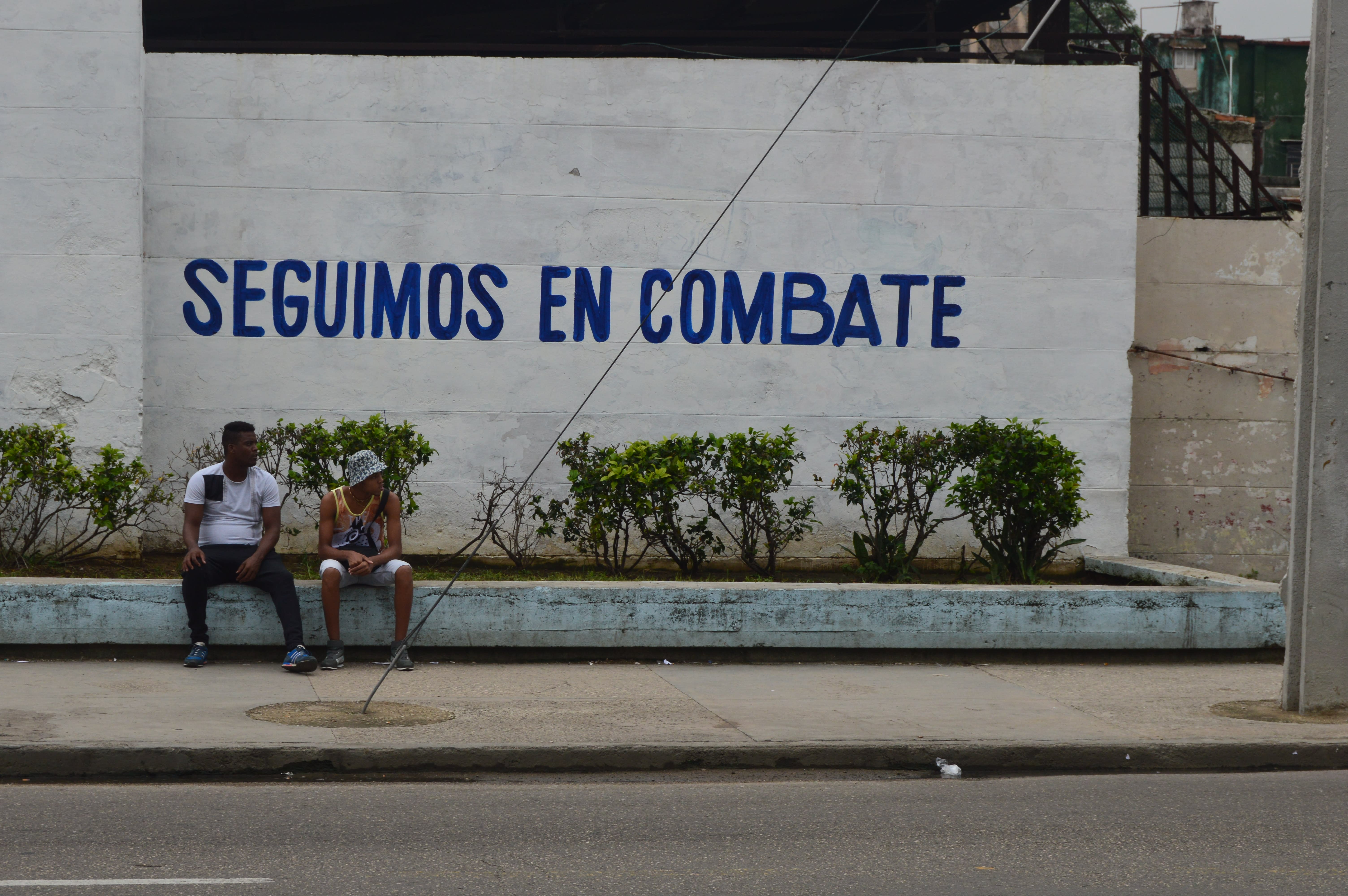 Dos personas sentadas frente a cartel revolucionario en La Habana: Seguimos en combate