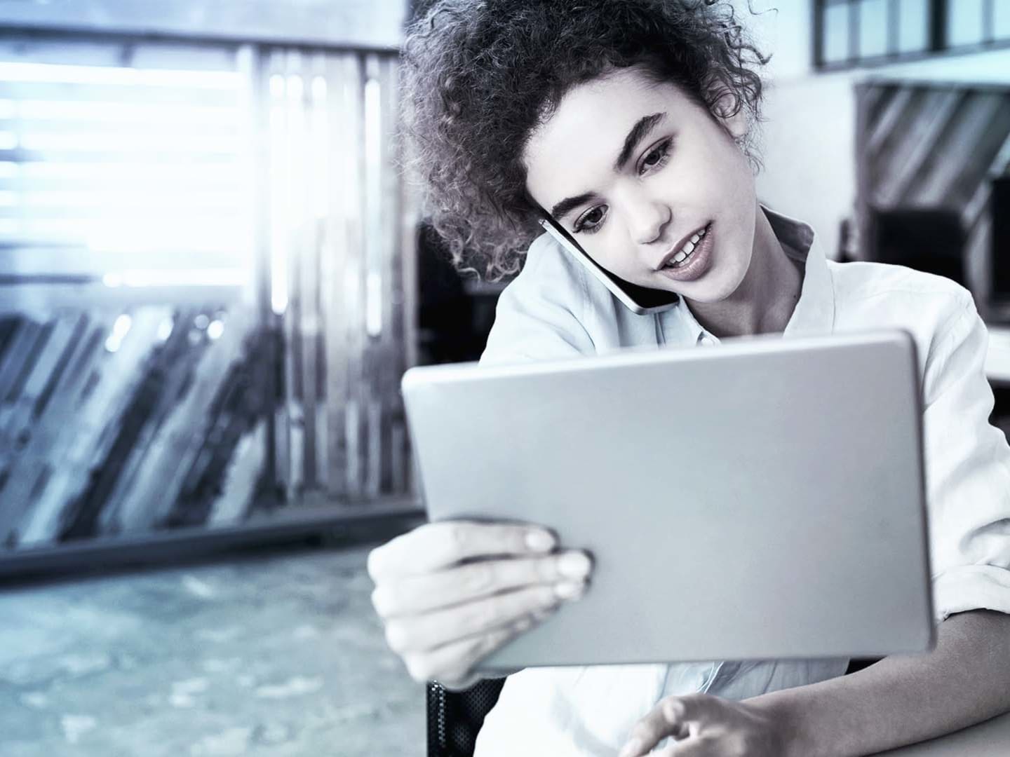 A young woman with curly hair is focused on a tablet device, with a smartphone cradled against her shoulder.