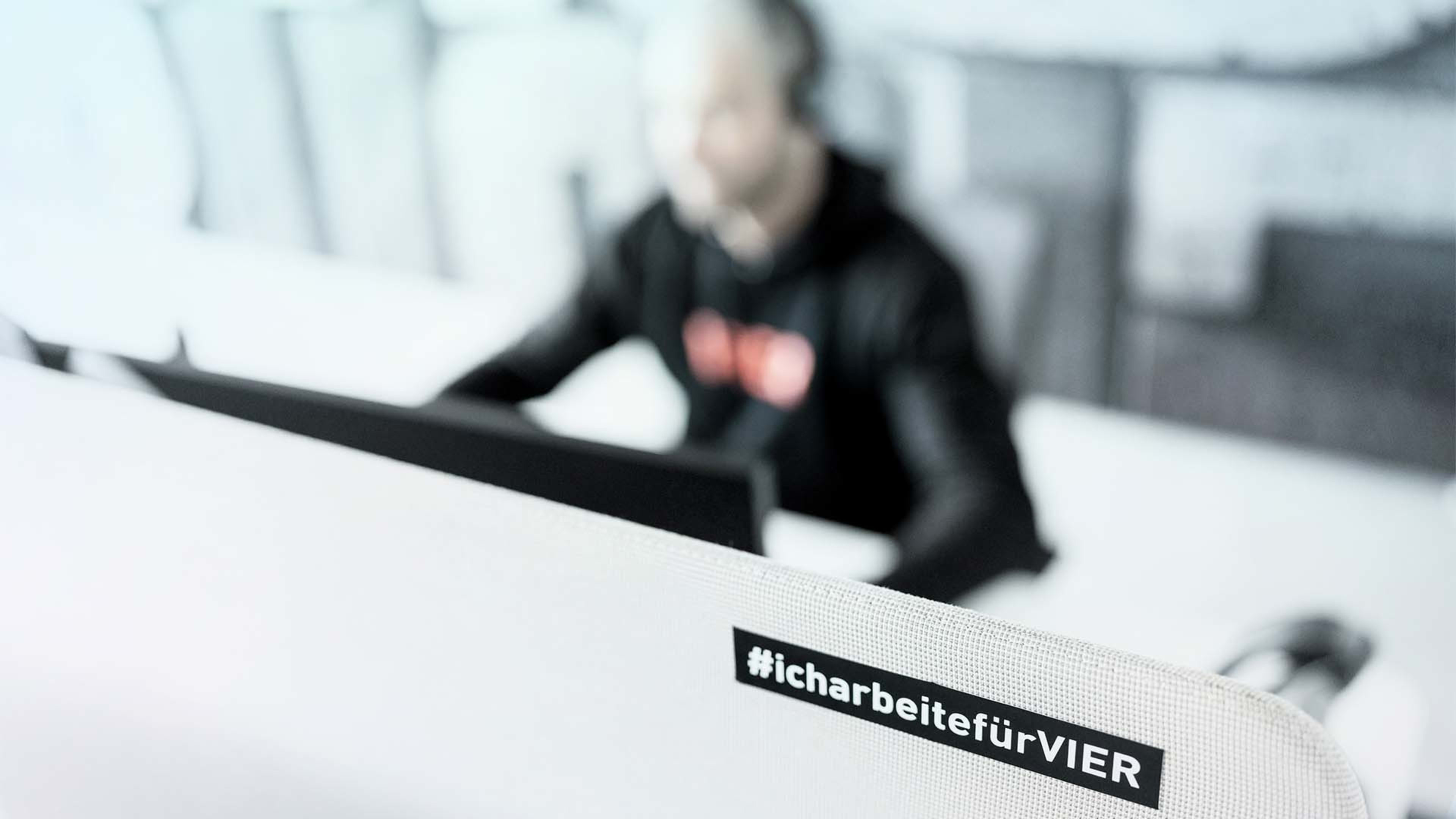 Sticker mit Hashtag #icharbeitefürVIER auf einem Raumtrenner, dahinter Person im VIER-Hoodie vor Bildschirm