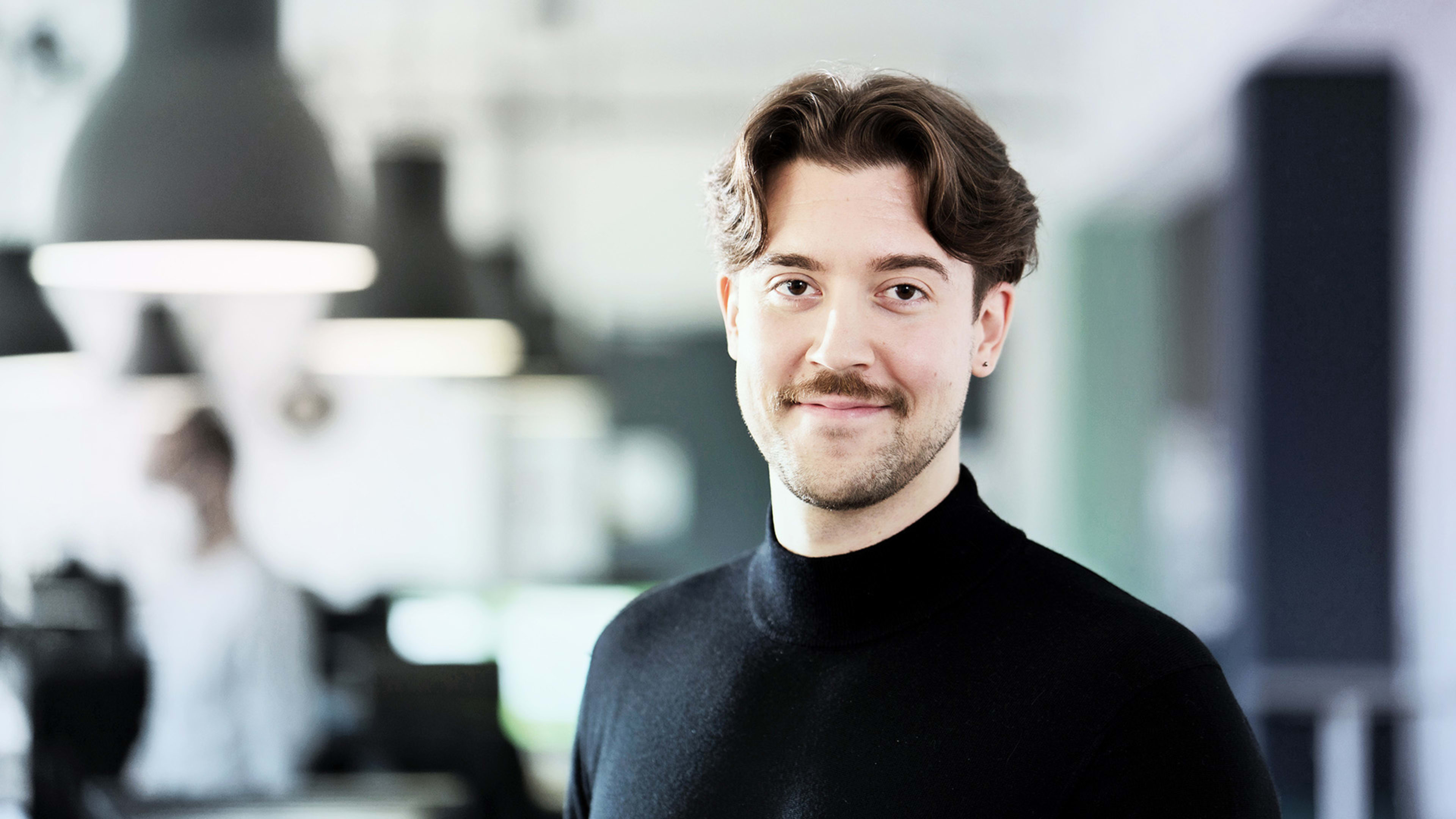 Lächelnder junger Mann mit braunem Haar, Bart und einem schwarzen Pullover, der in einem Büro steht.