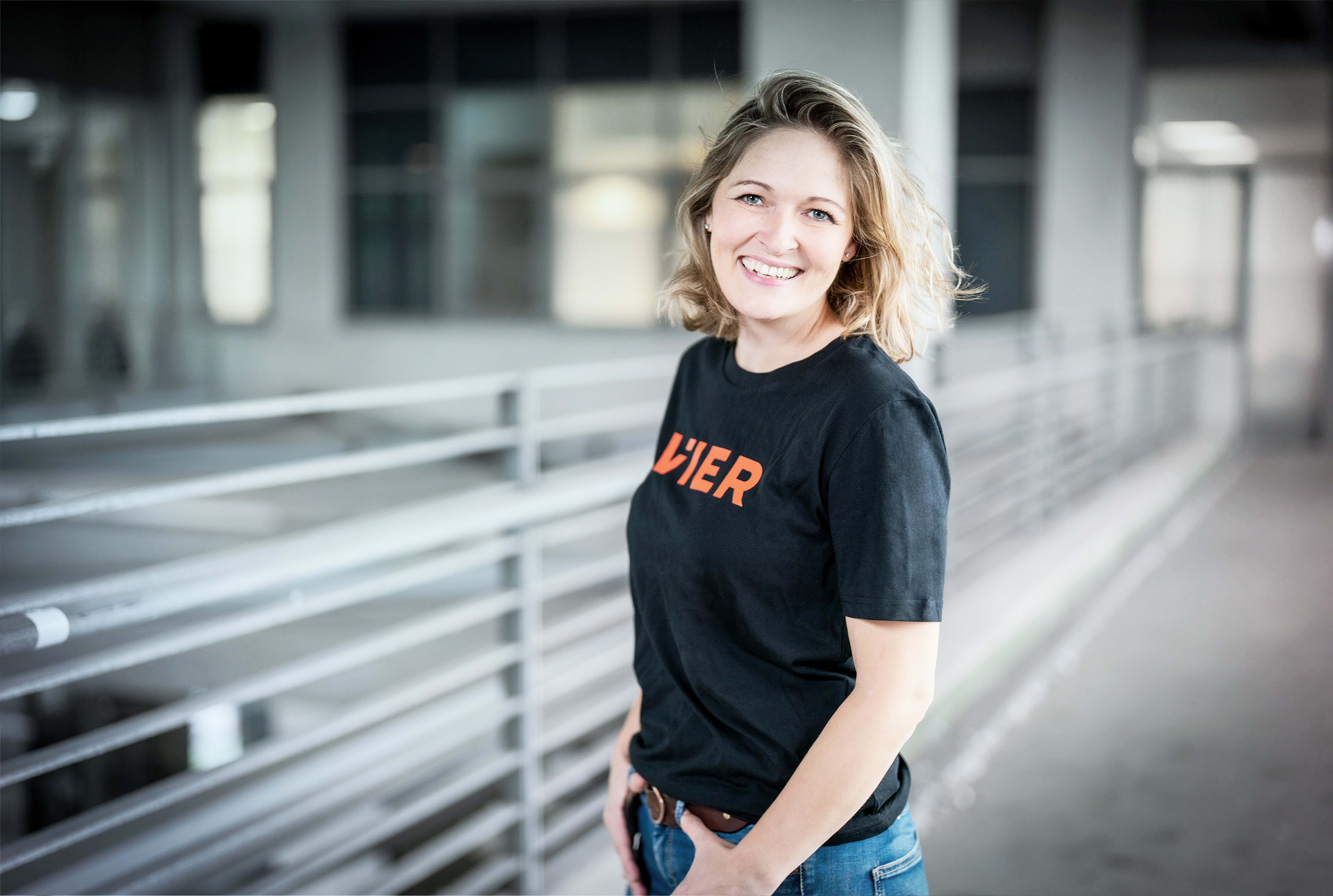 Frau mit blondem Haar, die ein schwarzes T-Shirt mit VIER Aufschrift trägt, steht vor einem grauen Gebäude und lächelt in die Kamera.