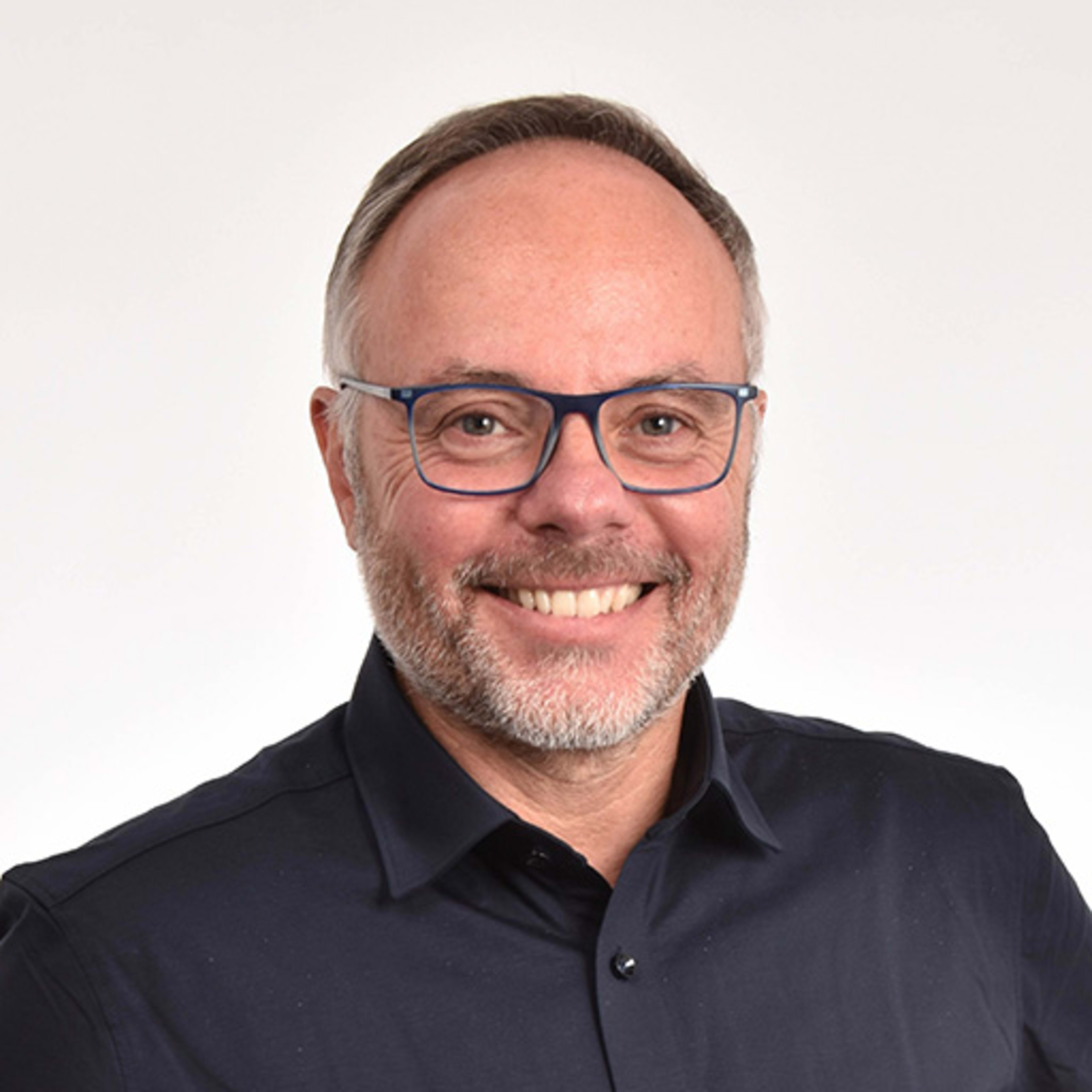 Ein lächelnder Mann mittleren Alters mit Brille und Bart, der ein dunkles Hemd trägt, vor einem weißen Hintergrund.