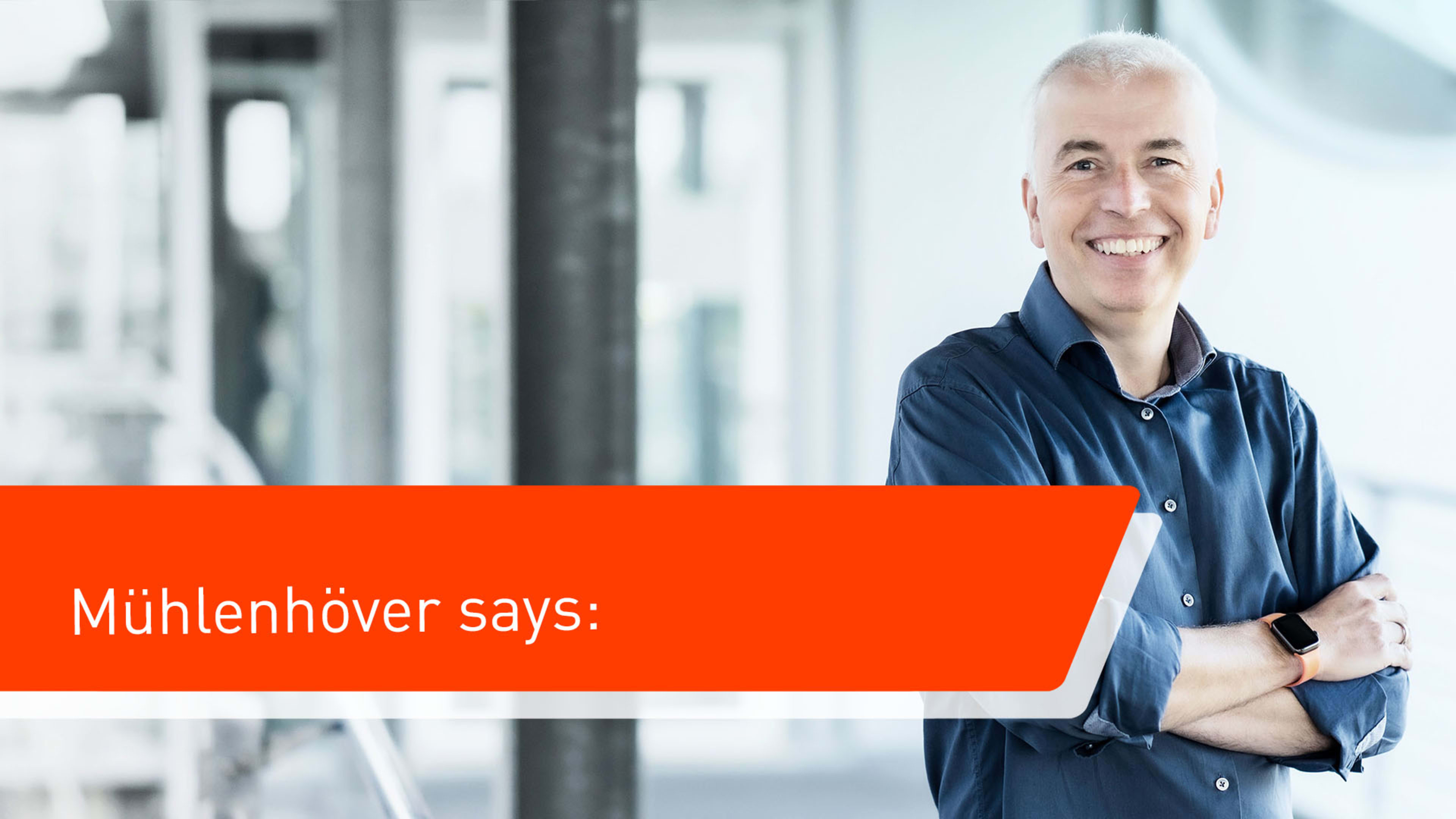 A smiling man with short white hair wearing a blue shirt and a smartwatch in front of an office building. An overlayed orange box says "Mühlenhover says".