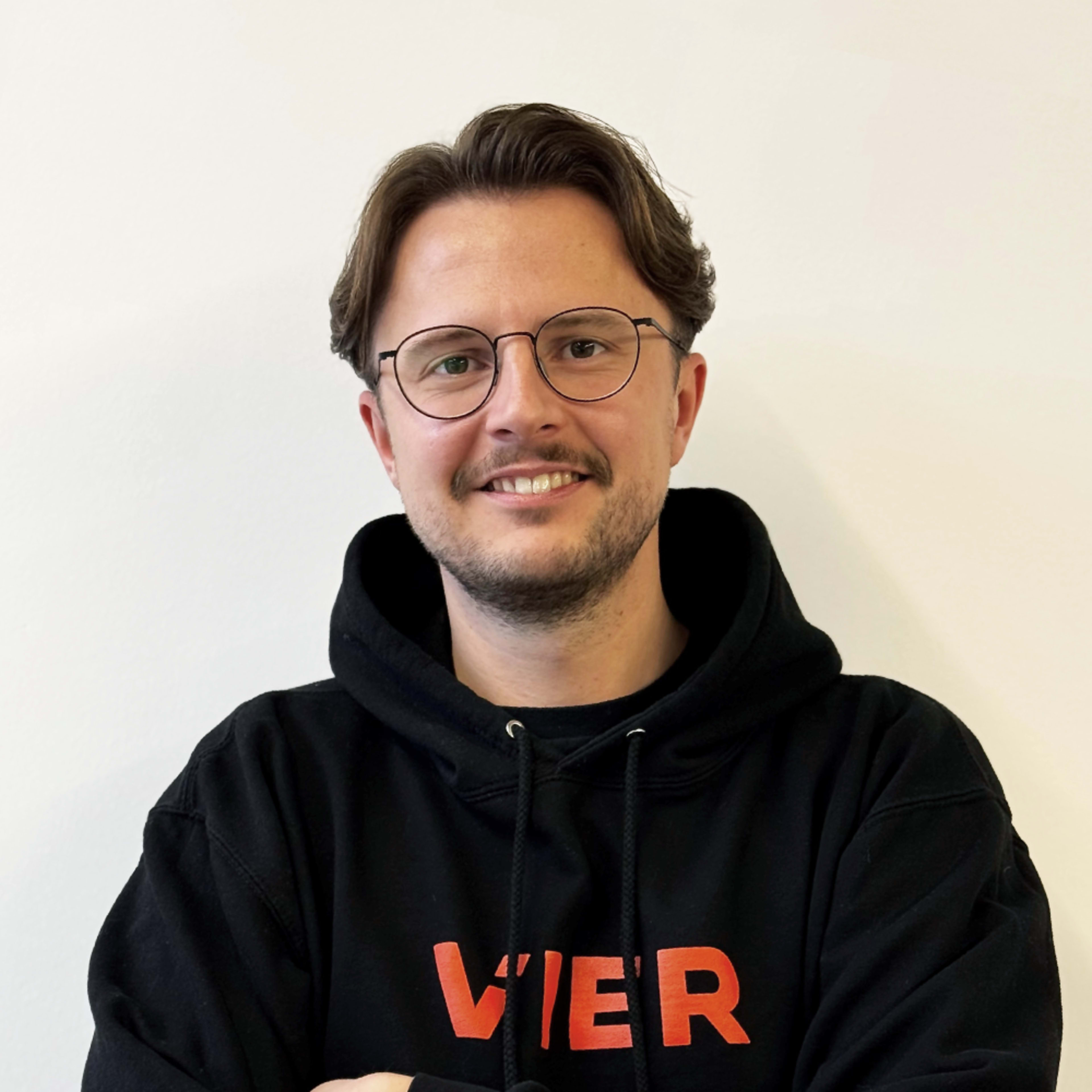 Ein lächelnder junger Mann mit Brille und einem schwarzen Kapuzenpulli mit der Aufschrift „VIER“ steht vor einem schlichten weißen Hintergrund.