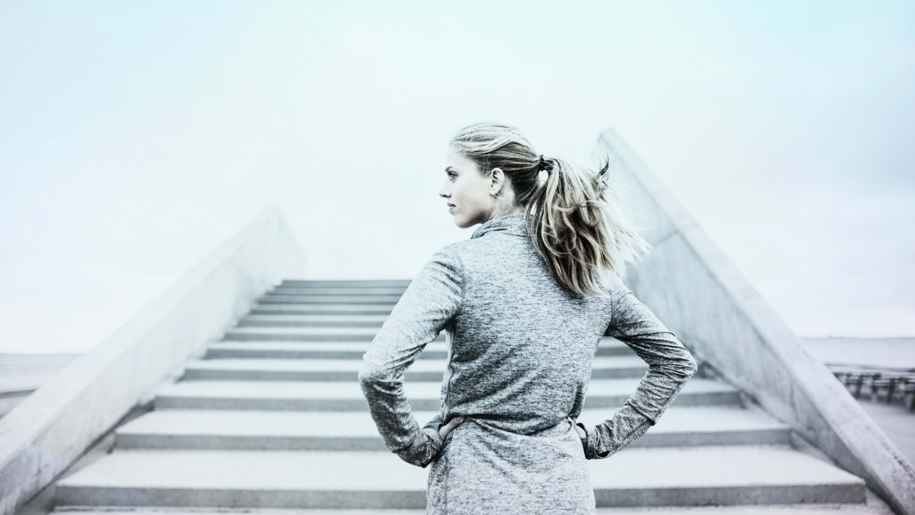 A woman with long hair stands on a staircase, her silhouette framed against a bright, hazy background.