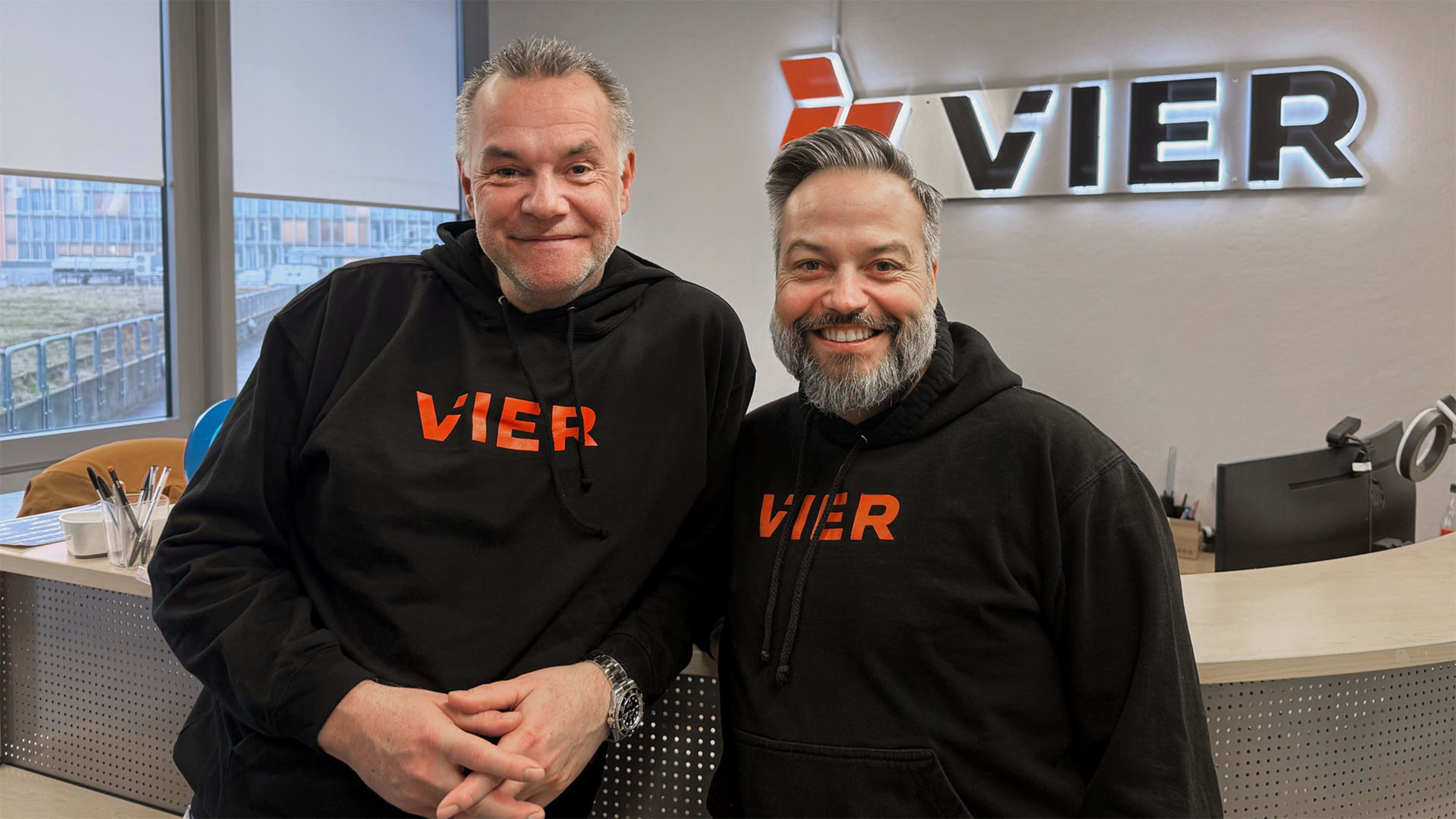 Zwei Männer in schwarzen Kapuzenpullovern mit dem Aufdruck „VIER“ stehen in einem Büro, in dem im Hintergrund ein großes „VIER“-Schild zu sehen ist.