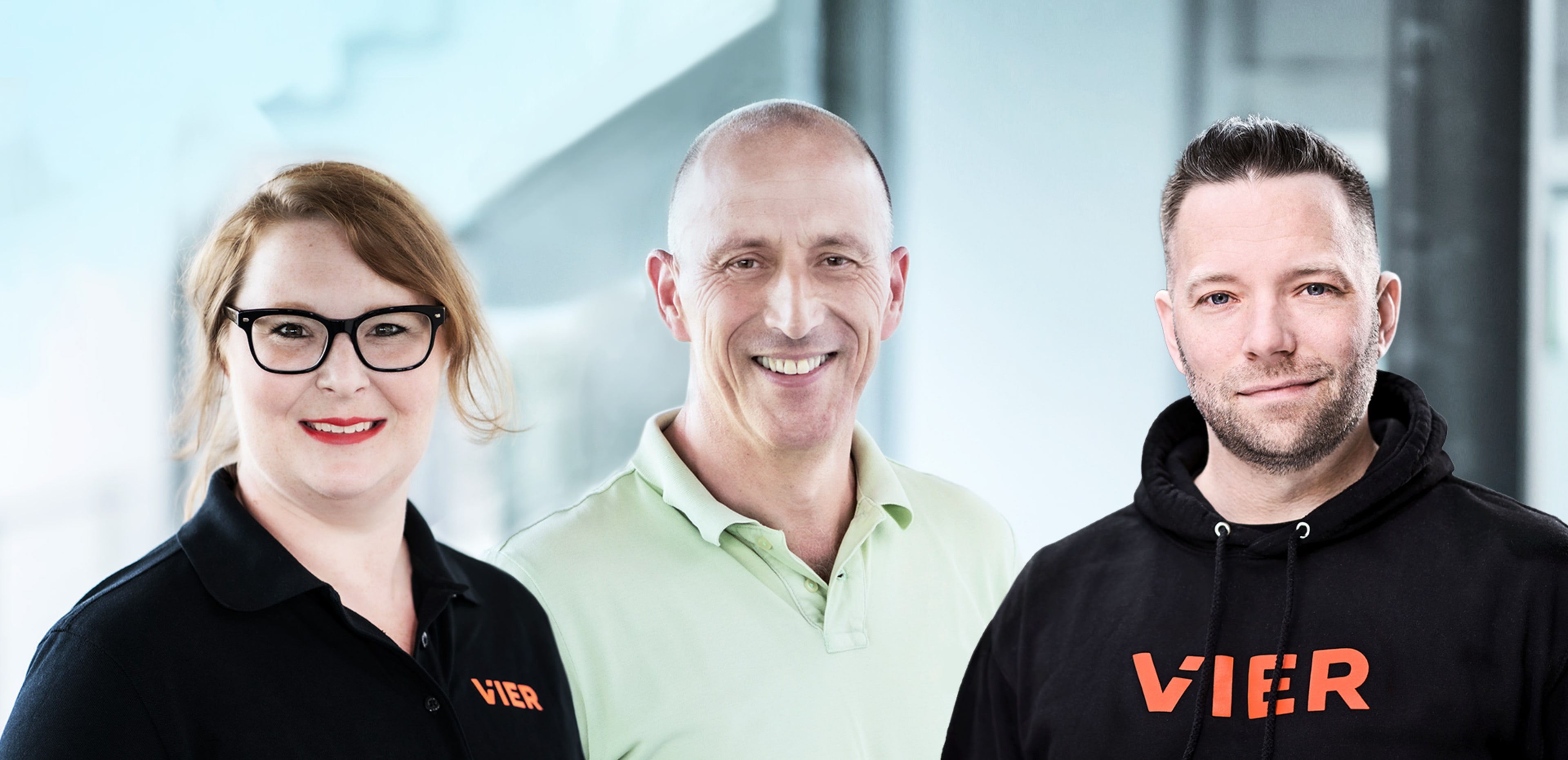 The image shows three people, two men and one woman, smiling and posing together in front of a blurred office building.