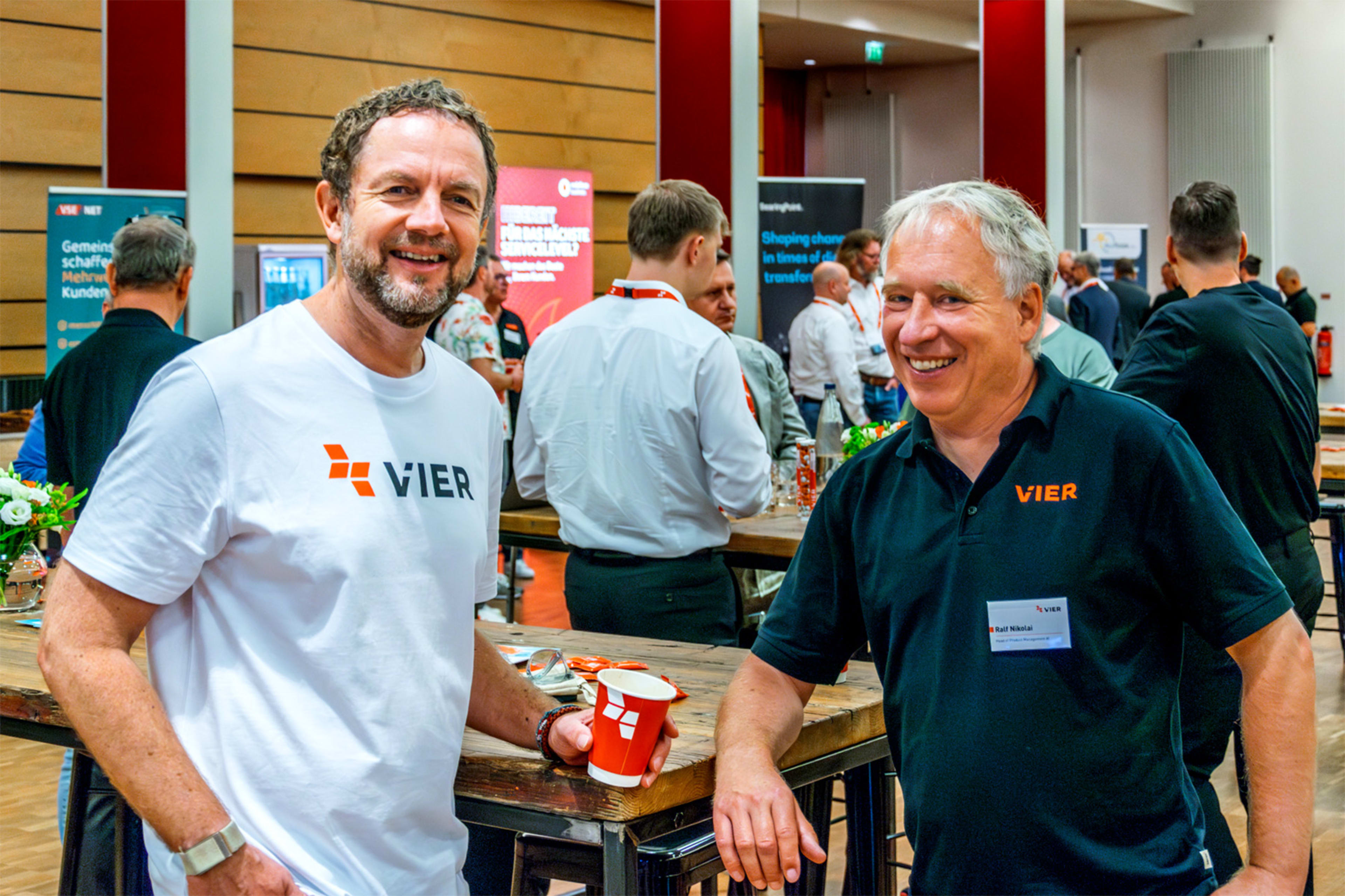 Das Bild zeigt Daniel Krantz, VIER Vice President AI, und Ralf Nikolai, VIER Head of Product Management AI, die lächelnd in einer Veranstaltungshalle stehen, während im Hintergrund weitere Personen zu sehen sind.