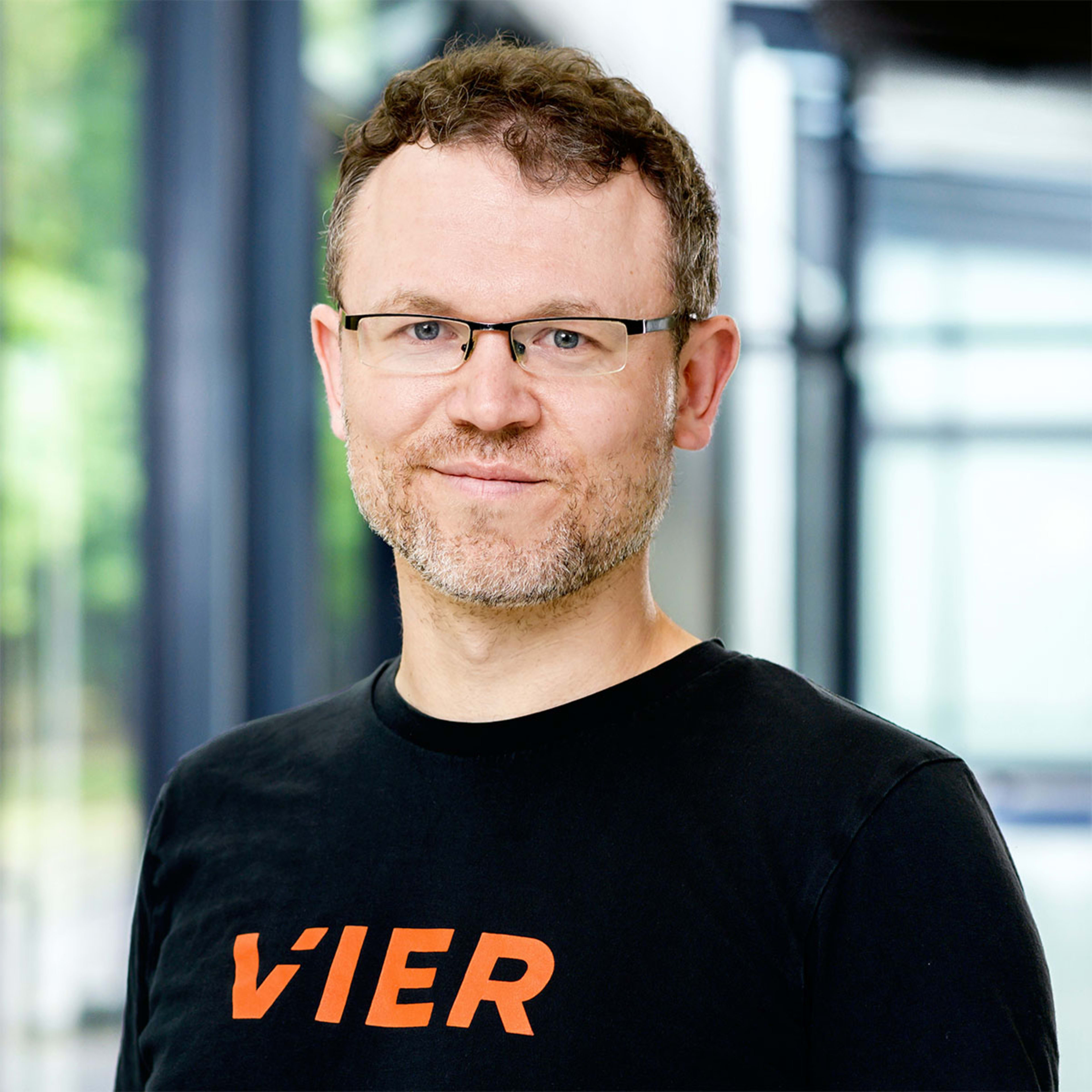 A middle-aged man with glasses and a beard wearing a black t-shirt with the word "VIER" printed on it, standing in front of a glass wall with a blurred outdoor scene in the background.