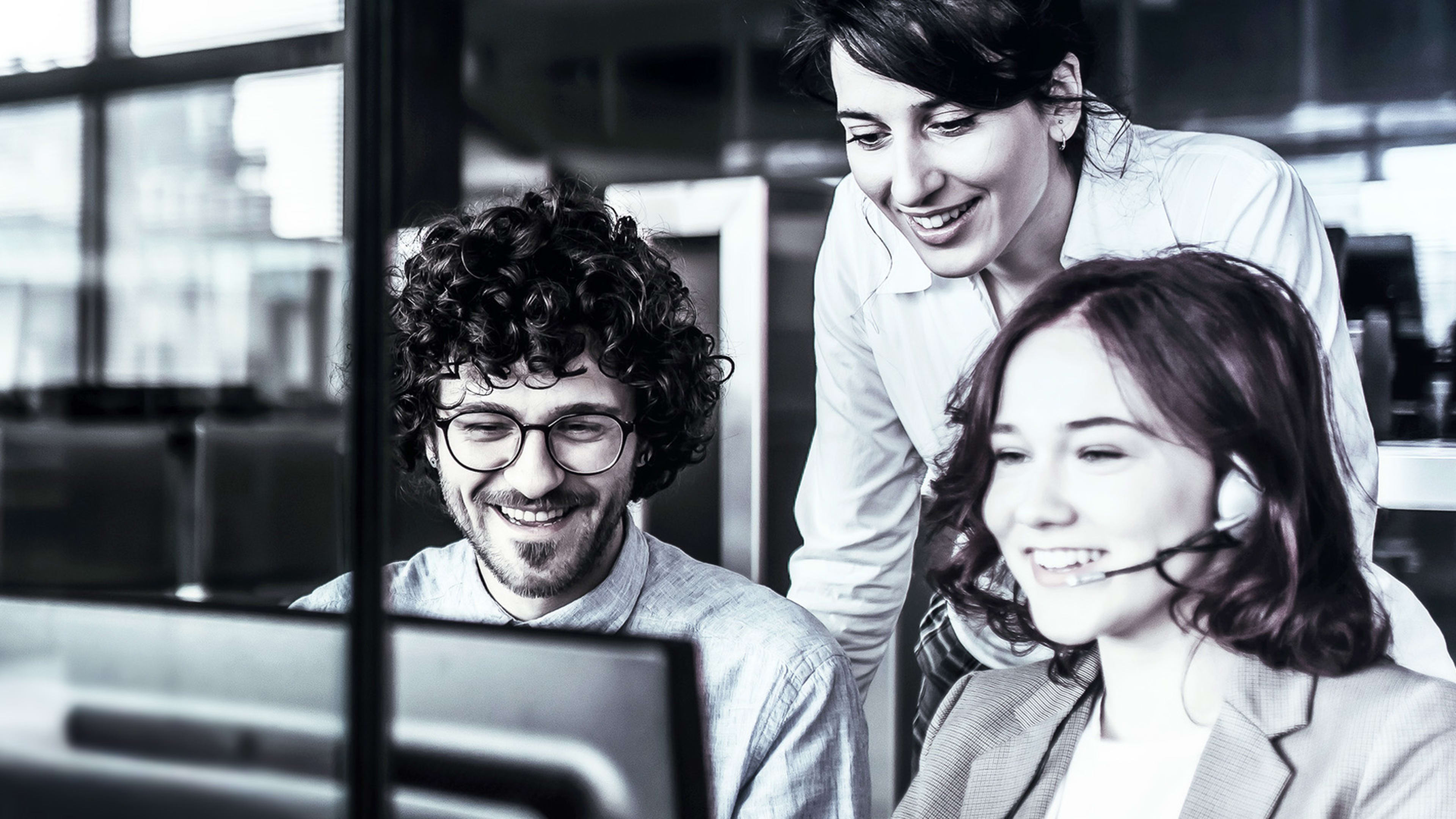 Ein Mann und zwei Frauen in einem Contact Center schauen lächelnd auf einen Desktop-Bildschirm.