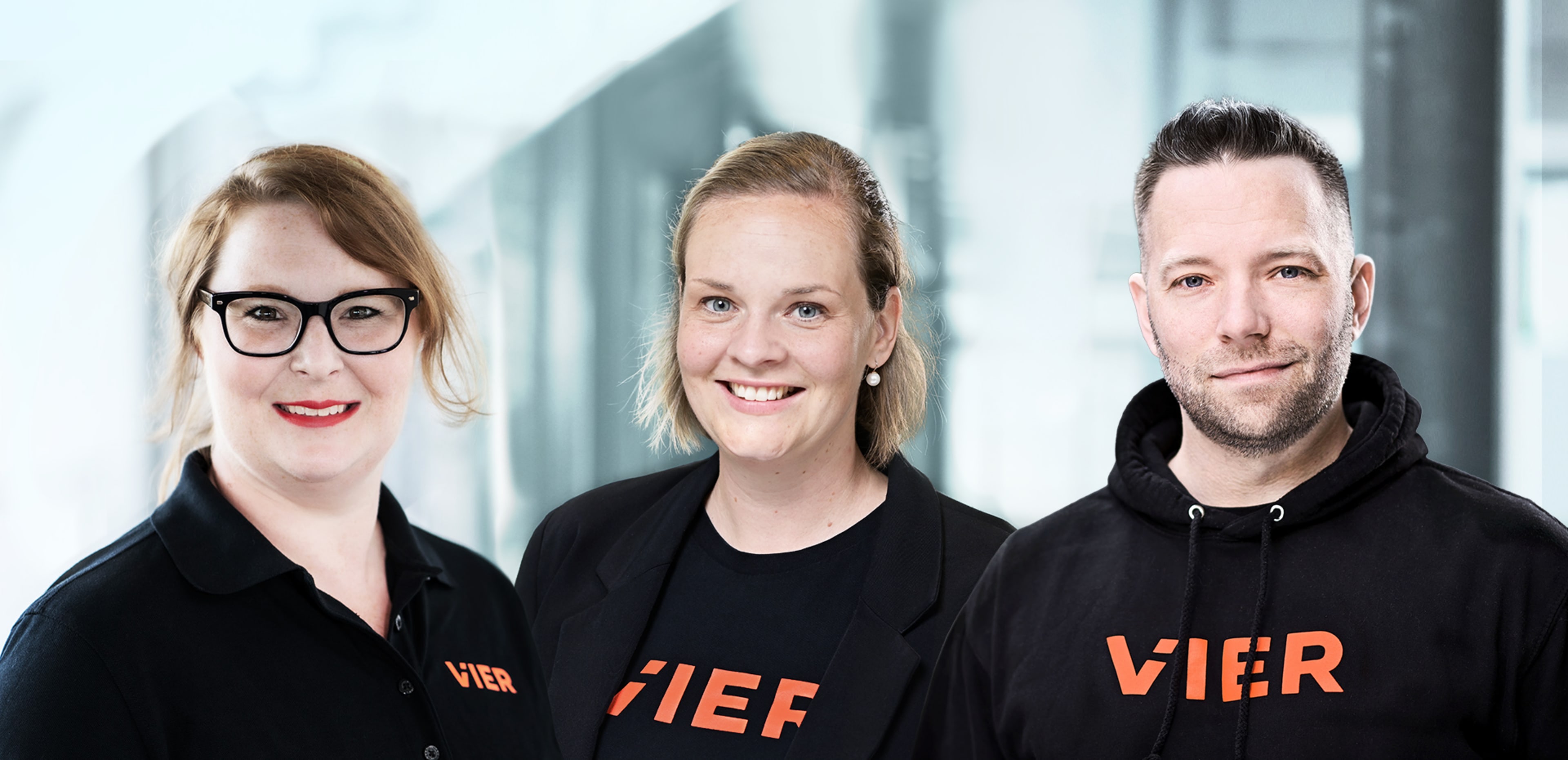 Drei Personen, zwei Frauen und ein Mann, stehen lächelnd vor einer unscharfen Büroumgebung.