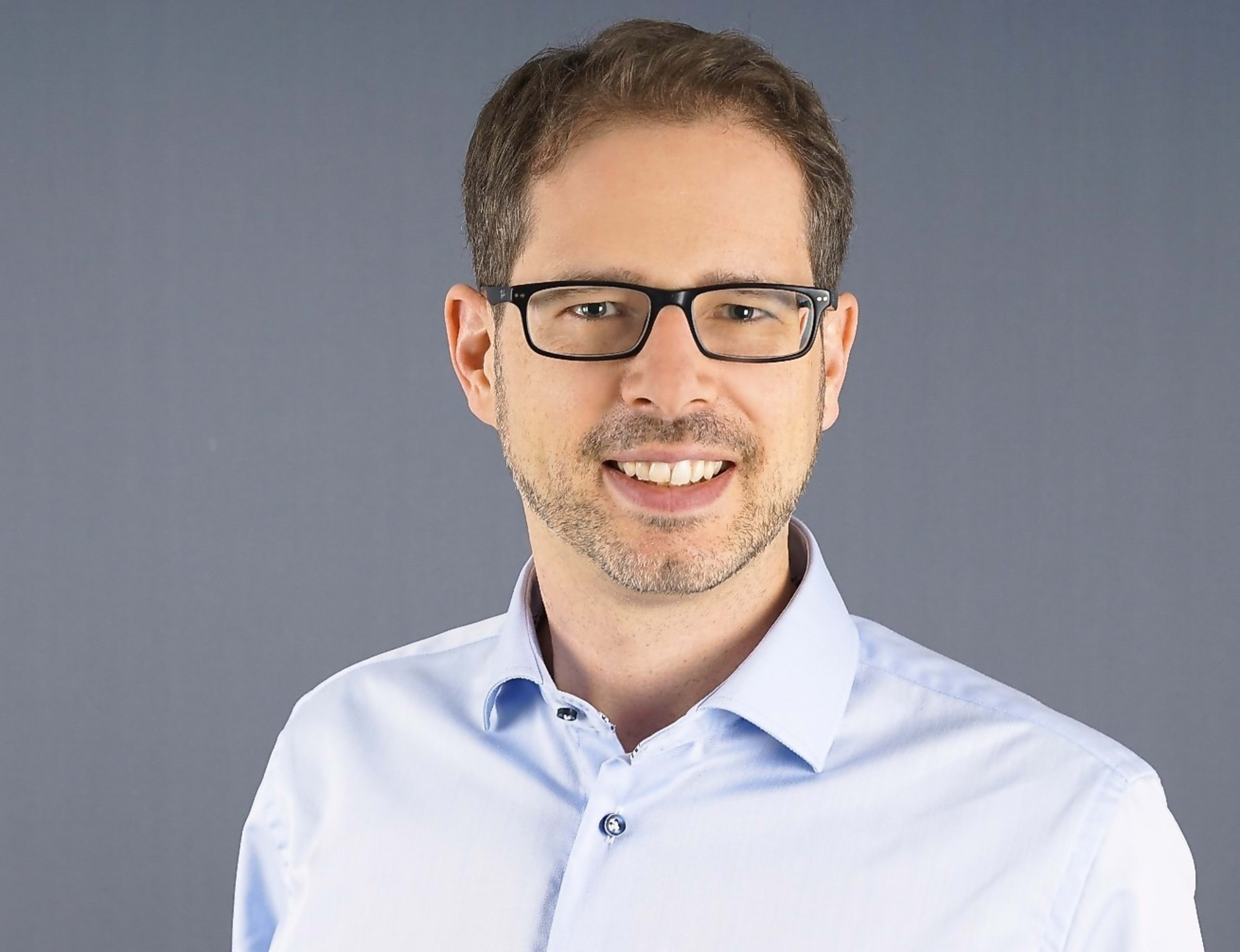 Ein lächelnder Mann mit Brille und Bart, bekleidet mit einem hellblauen Hemd, vor grauem Hintergrund.