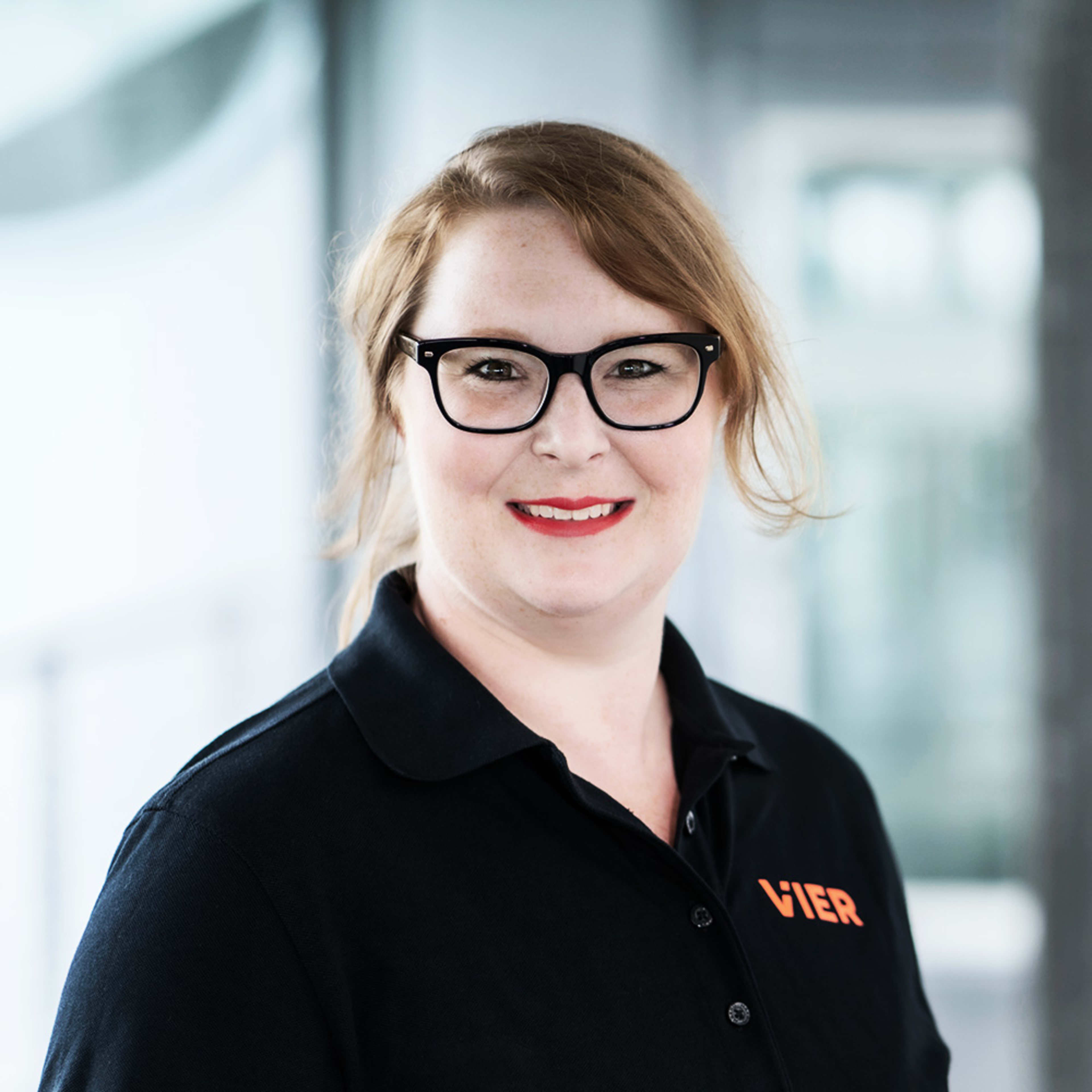Portrait of a woman with a friendly smile, reddish-blonde hair, and black glasses, wearing a black polo shirt with the word “VIER” printed on it.