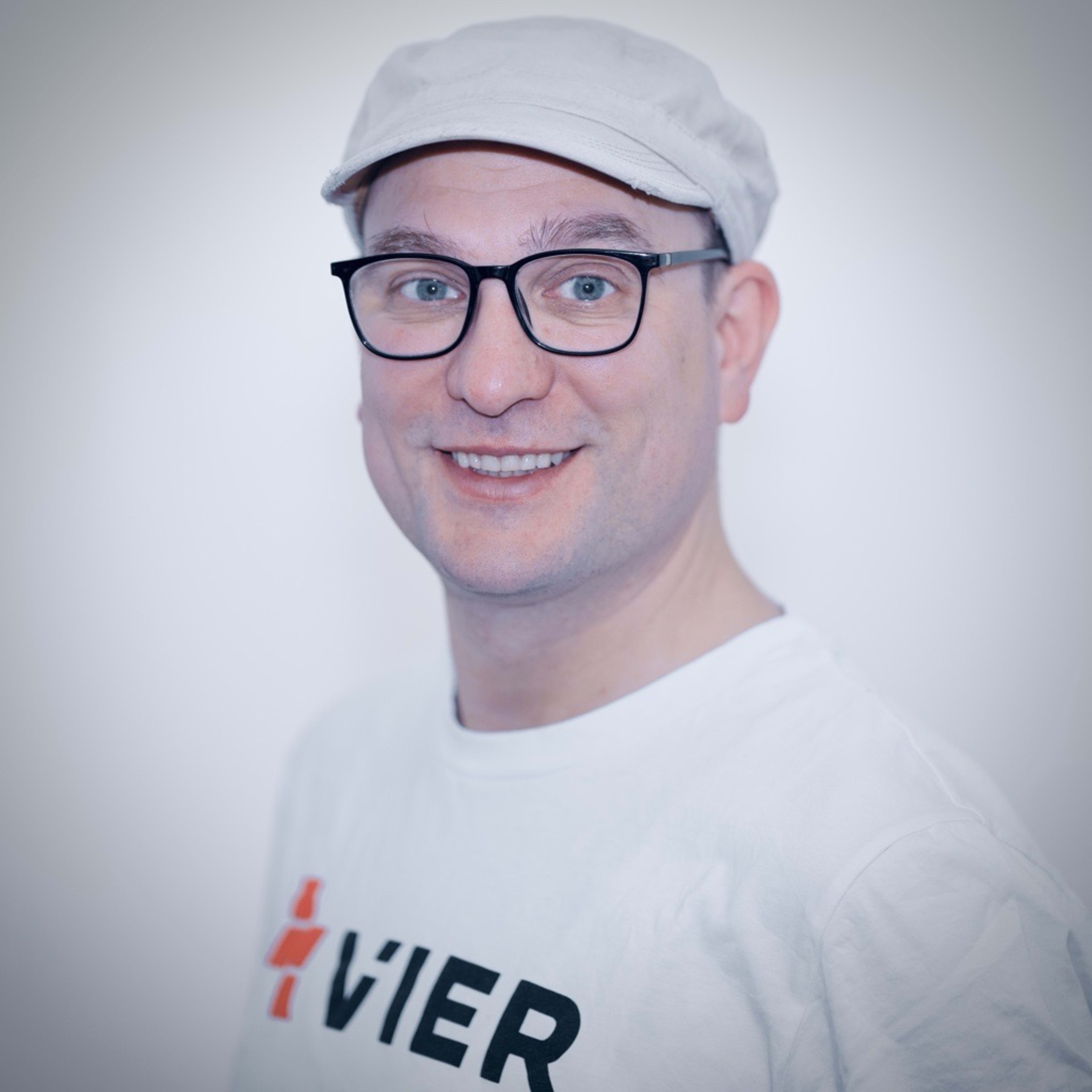 Ein Mann mit einer hellen Mütze und einer Brille lächelt vor einem schlichten weißen Hintergrund in die Kamera.