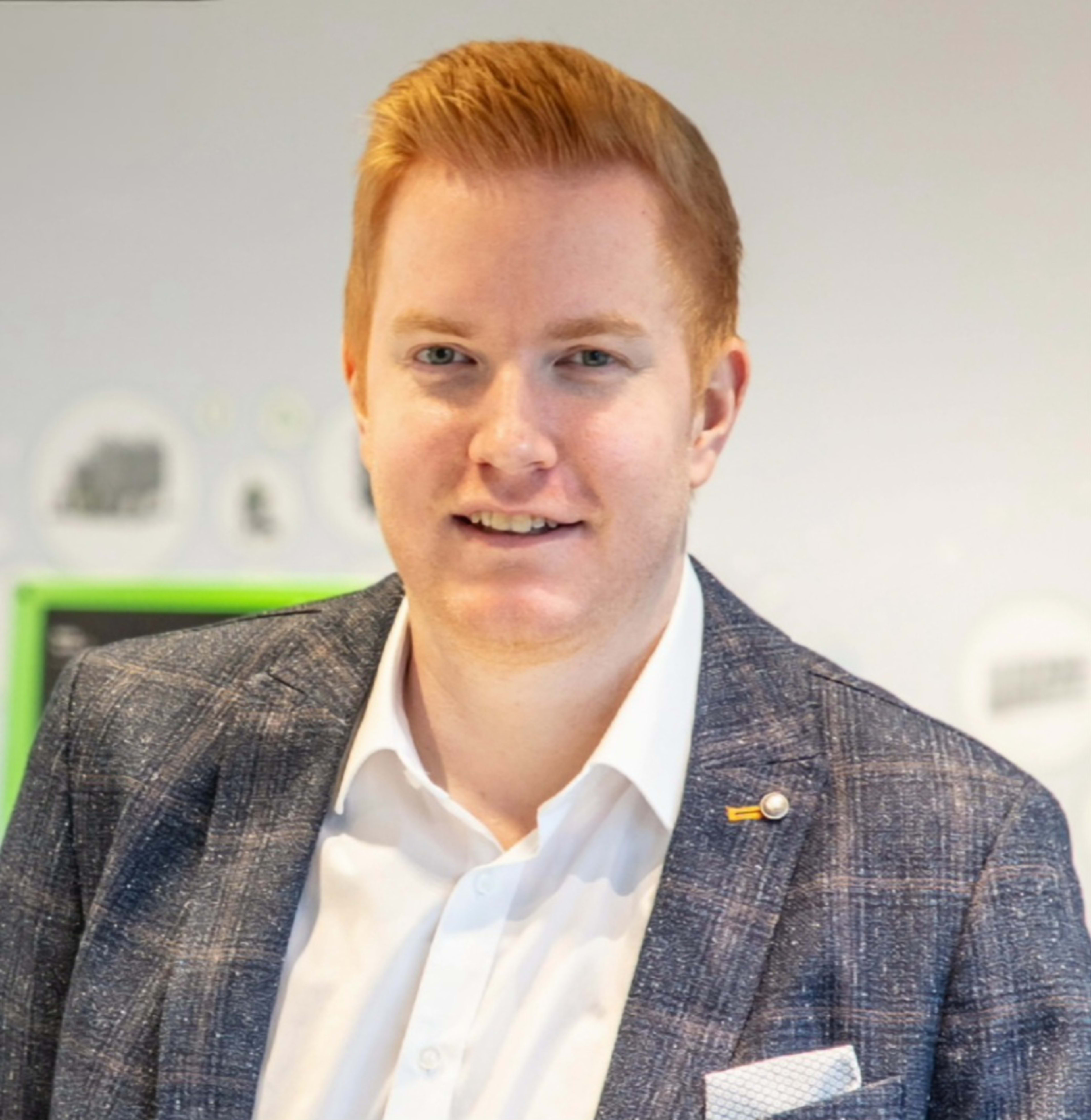 A man with red hair and a checkered jacket smiles at the camera in front of a blurred background.