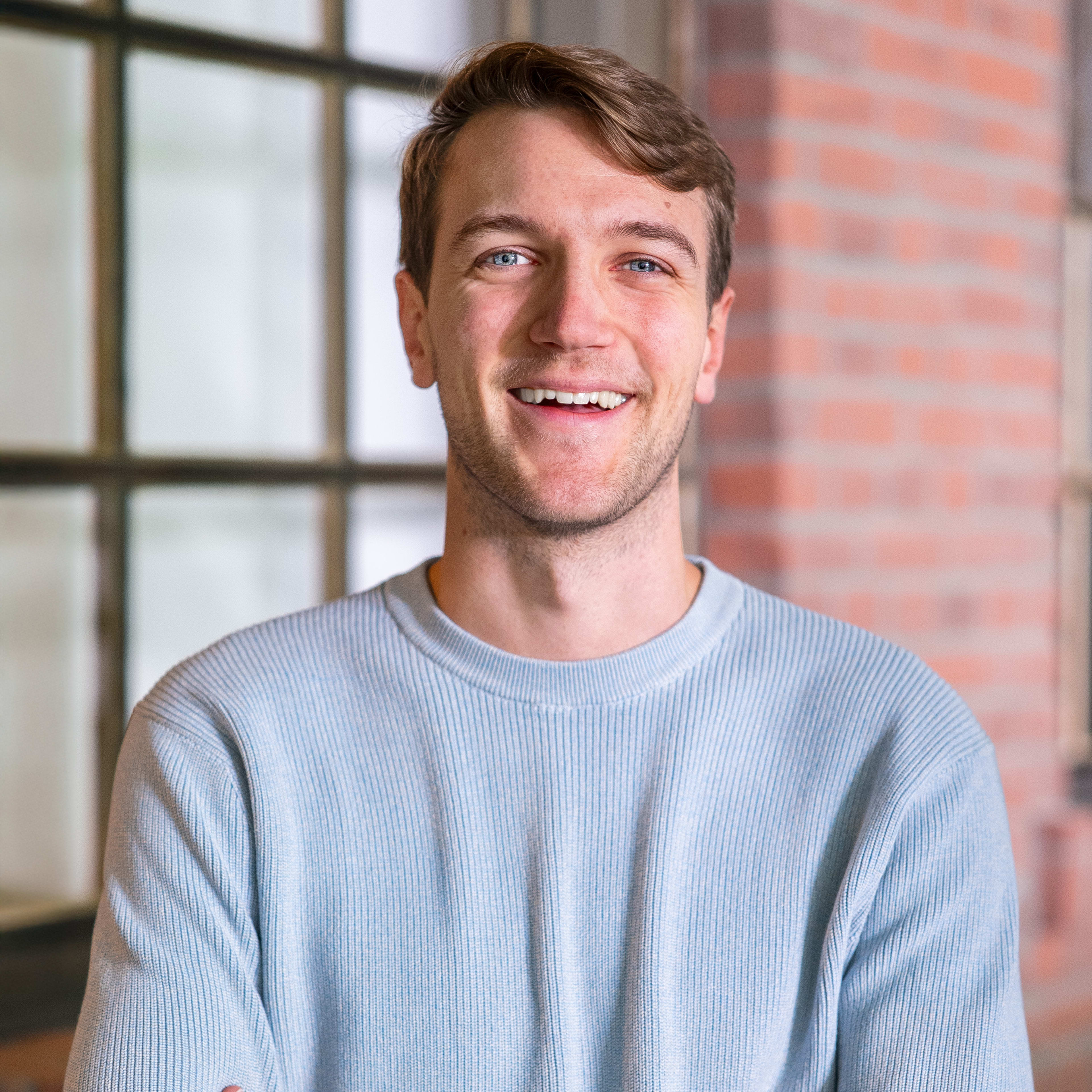A smiling man with brown hair and a light blue sweater standing in front of a blurred background of a window and brick wall.