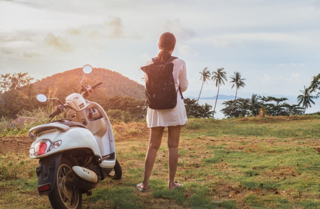frau-mit-rucksack-blickt-auf-das-meer-neben-ihr-steht-ein-motorroller.jpeg