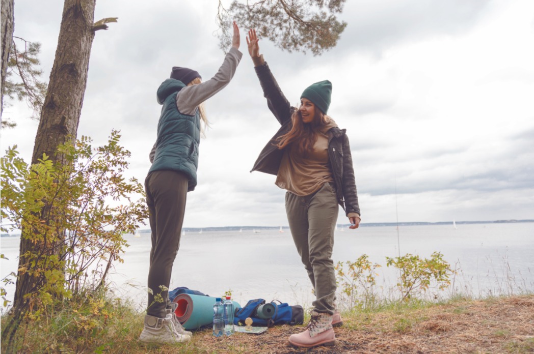 zwei-frauen-stehen-an-einem-see-geben-sich-ein-high-five-am-boden-liegen-campingutensilien.jpeg