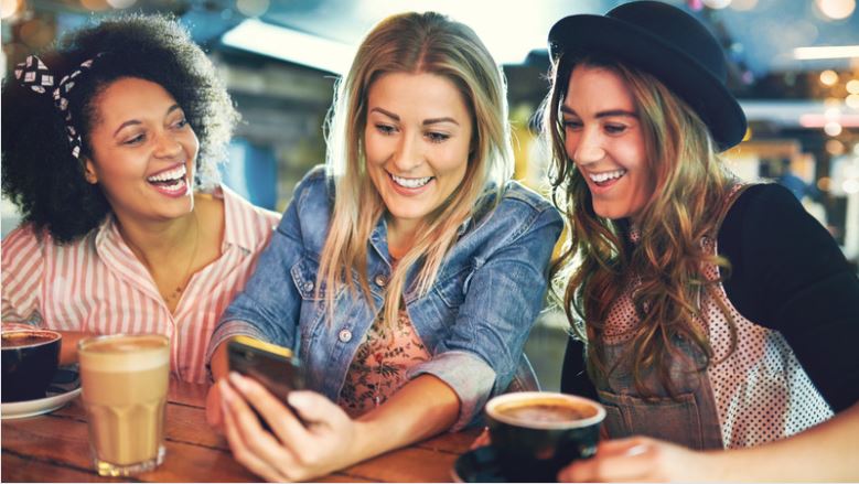 drei-frauen-sitzen-in-einem-cafe-und-schauen-laechelnd-auf-ein-smartphone.jpeg