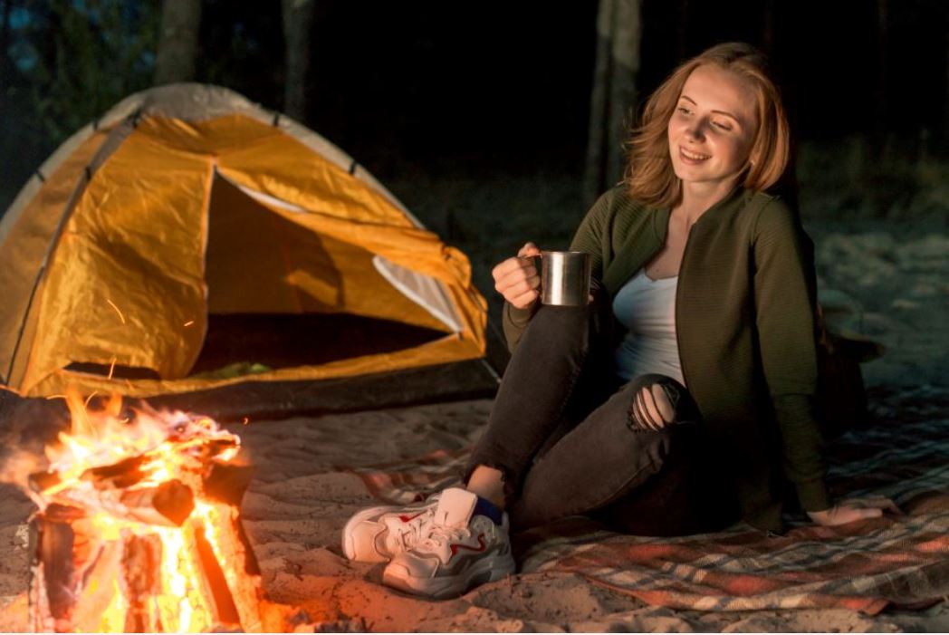 frau-sitzt-auf-einer-decke-vor-einem-lagerfeuer-und-hat-eine-tasse-in-der-hand-neben-ihr-steht-ein-zelt.JPG