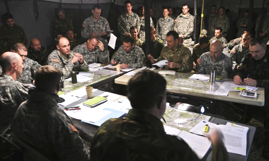 Group of military personnel in uniform gathered around a table during a strategic planning session. Maps, documents, and other planning materials are spread across the table, with officers discussing tactics. The image illustrates a wargaming or military strategy meeting, emphasizing teamwork and decision-making in a tactical environment.
