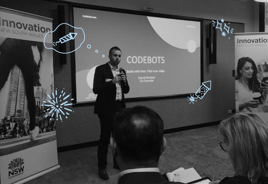 A photo of a man presewnting to a group of people. He is standing infron od a screen with the words CODEBOTS on it 