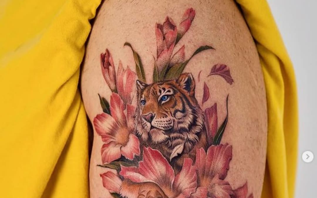 A tiger among pink blossoms, a colour fine-line tattoo on an upper arm by Nando.