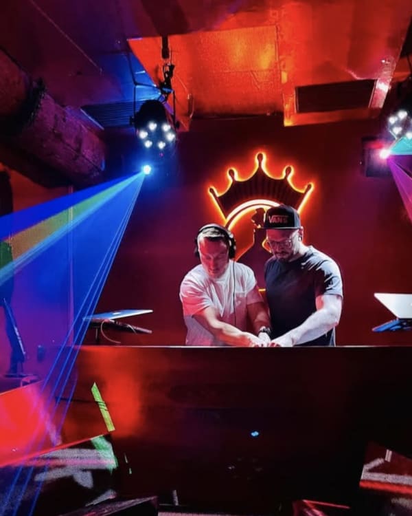 Two DJs play side by side in a club booth, lit by laser beams under a neon crown.