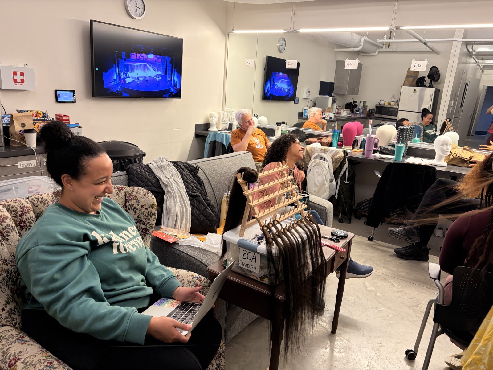 The hair station setup at Umbrella Arts Center's Hairspray production, with wig stands, named station tags for each performer, and the production's stage feed visible on the monitor.