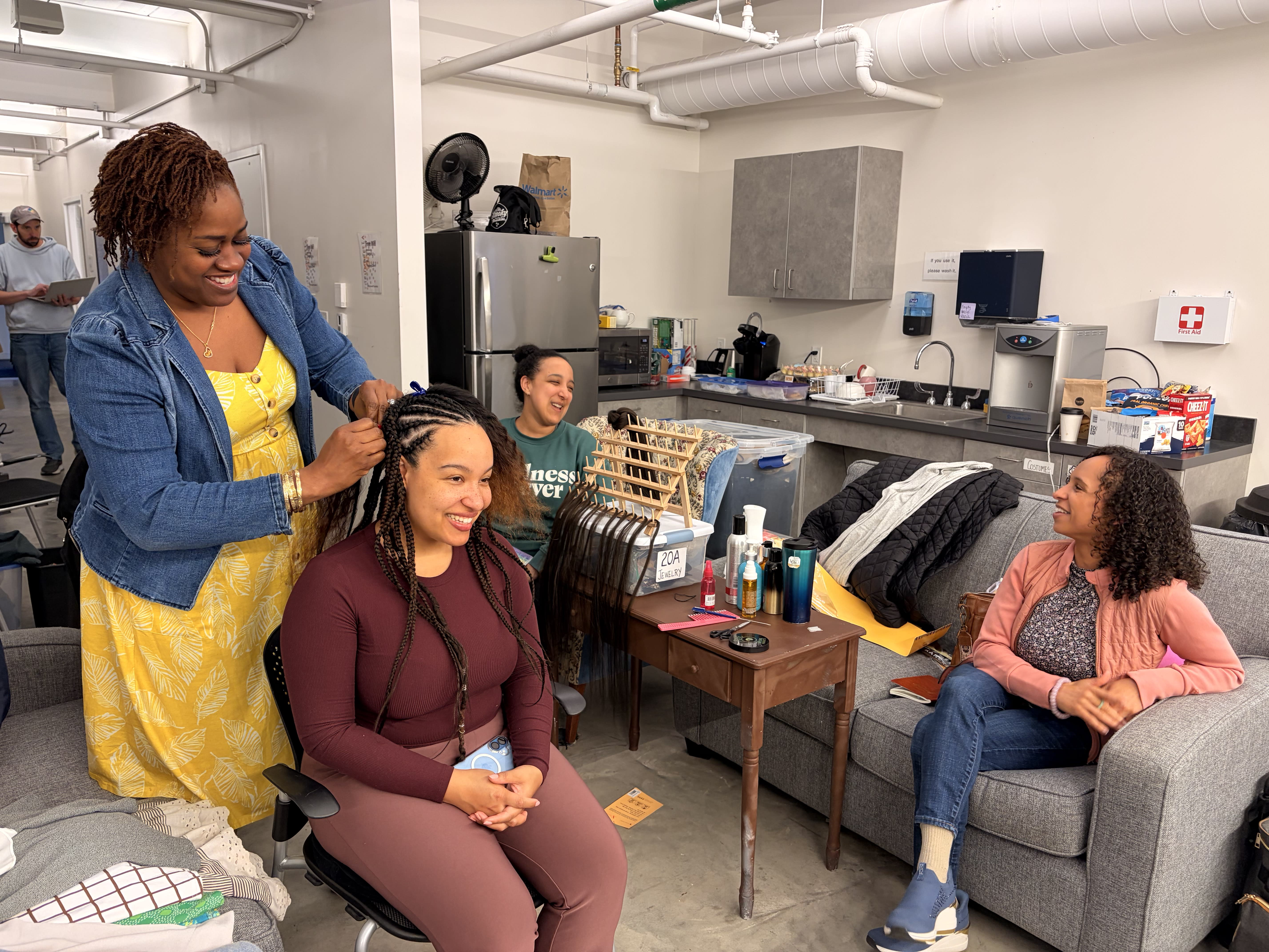 A qualified Hair Lead from the Texture Ready team braiding a performer's protective style at the Umbrella Arts Center production of Hairspray, with Kira Troilo observing.