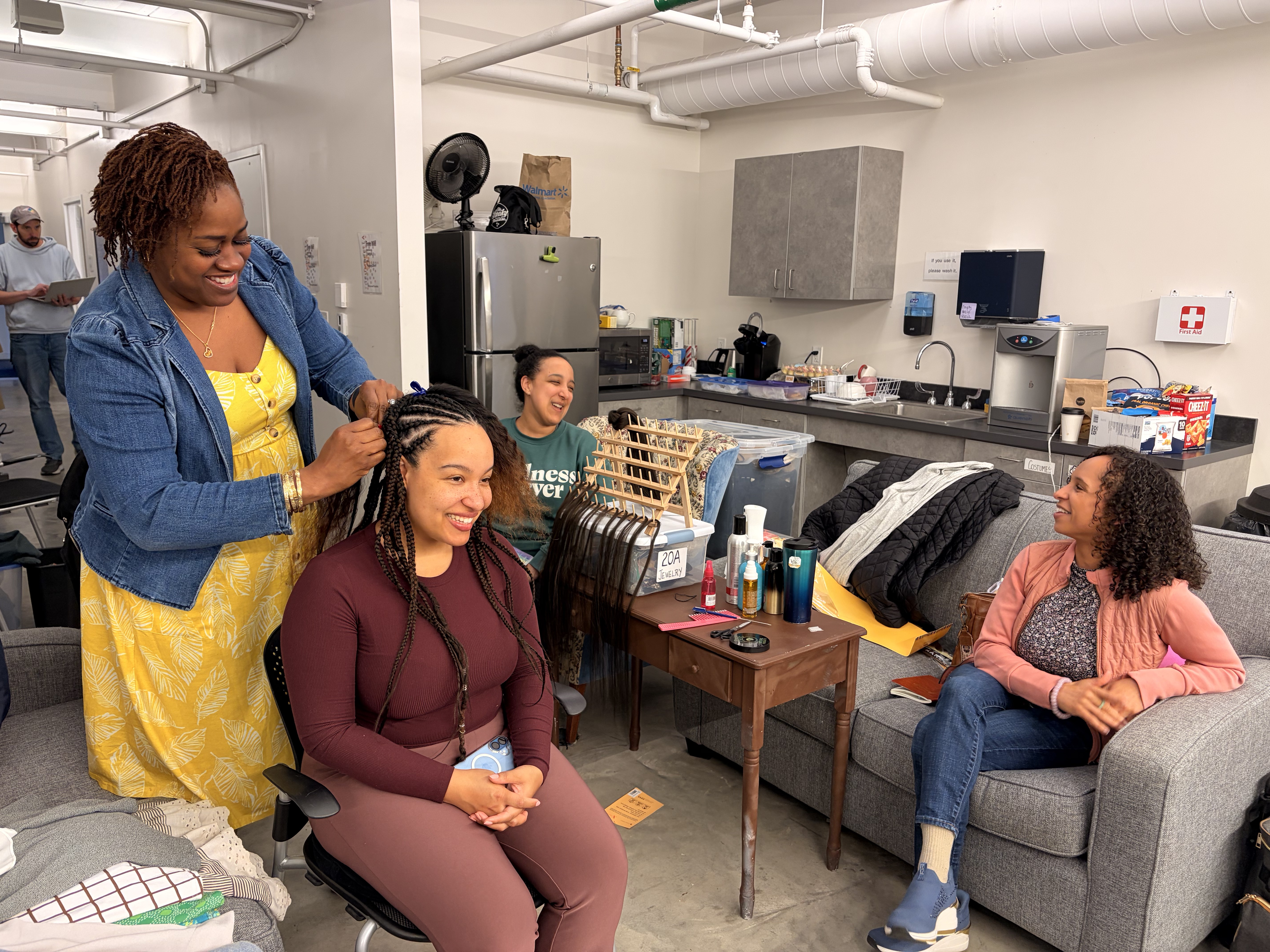 Texture Ready Salon Day in a green room at the Umbrella Arts Center: a braider installs a protective style on a seated performer while castmates talk nearby