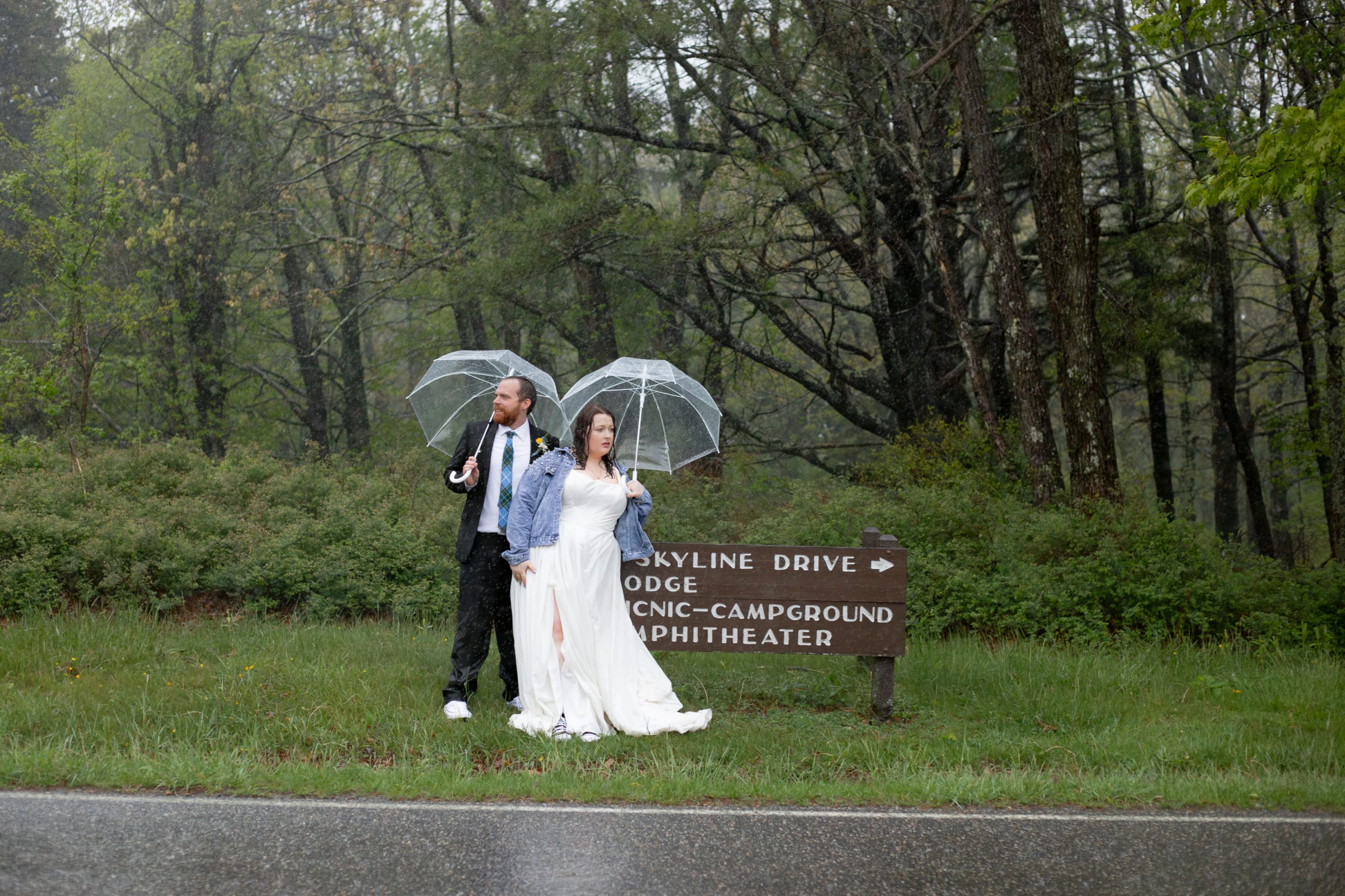 Couple standing next to a Skyline Drive sign in the rain, each holding a clear bubble umbrella, bride in a denim jacket and Converse sneakers