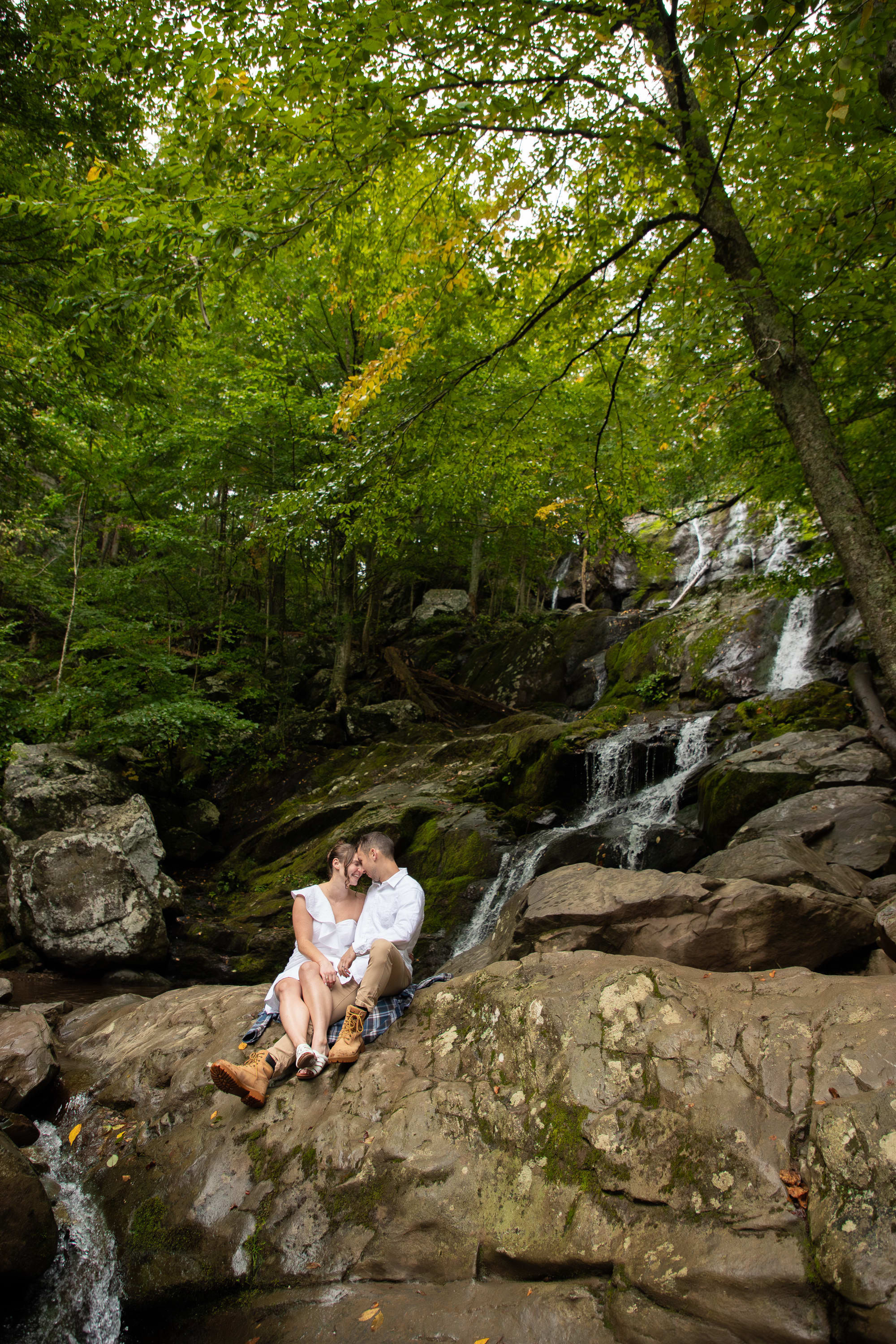 Couple sitting on moss-covered rocks beneath a lush green canopy beside a waterfall in Shenandoah National Park