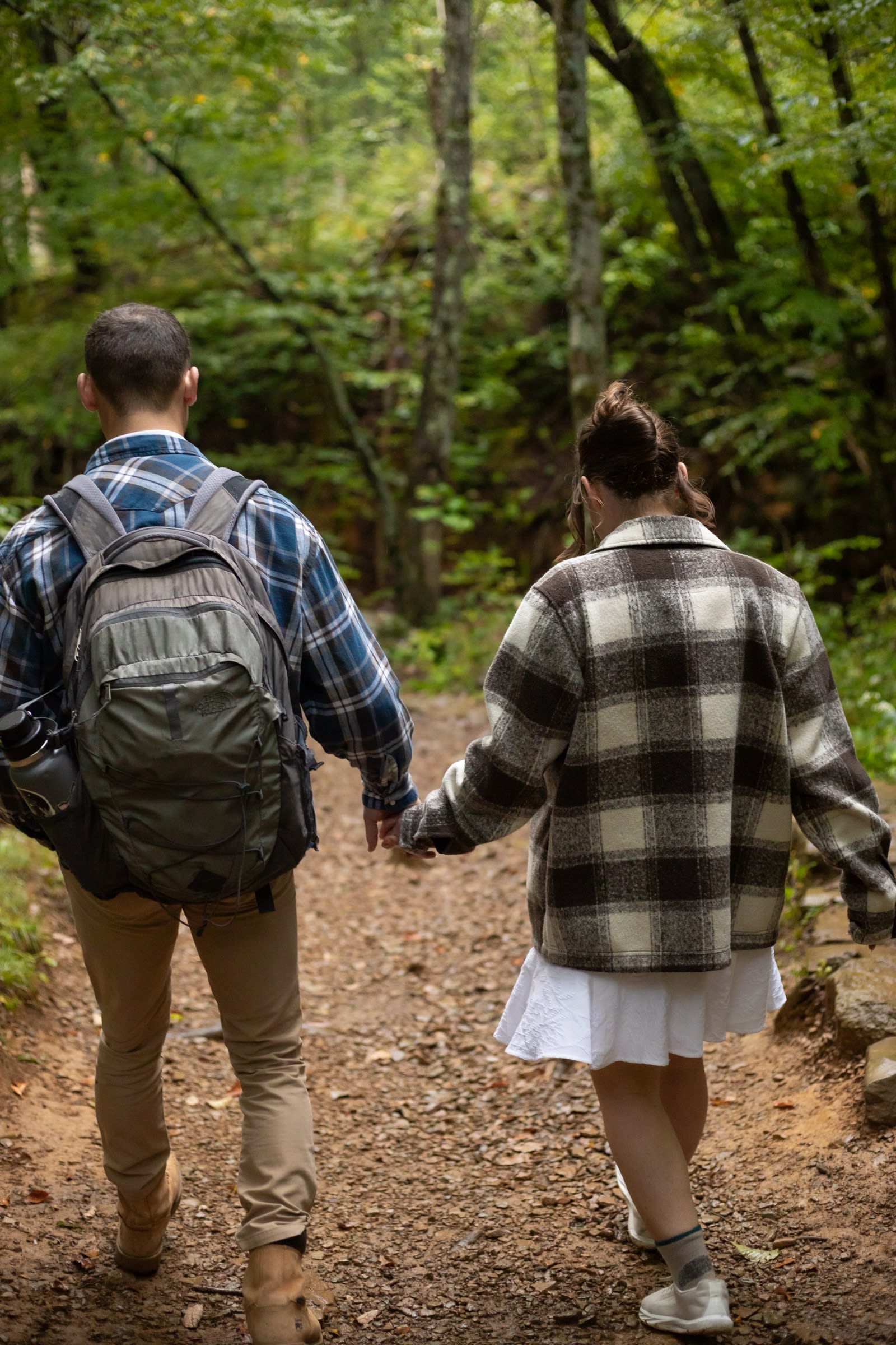 Couple hiking hand in hand on a green forest trail in Shenandoah National Park, one carrying a backpack