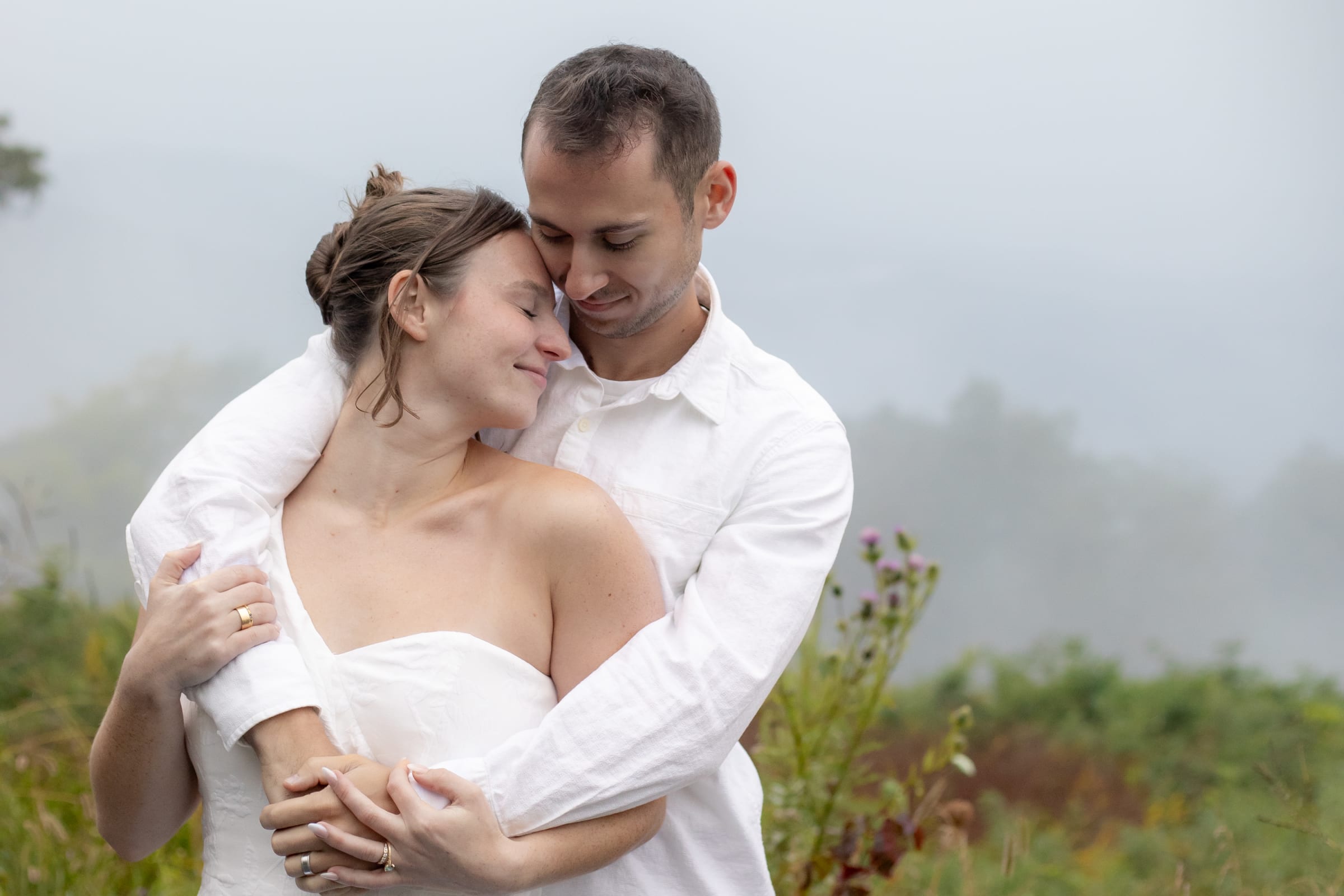 Couple embracing in the fog at Shenandoah National Park, soft overcast light wrapping around them in a misty meadow